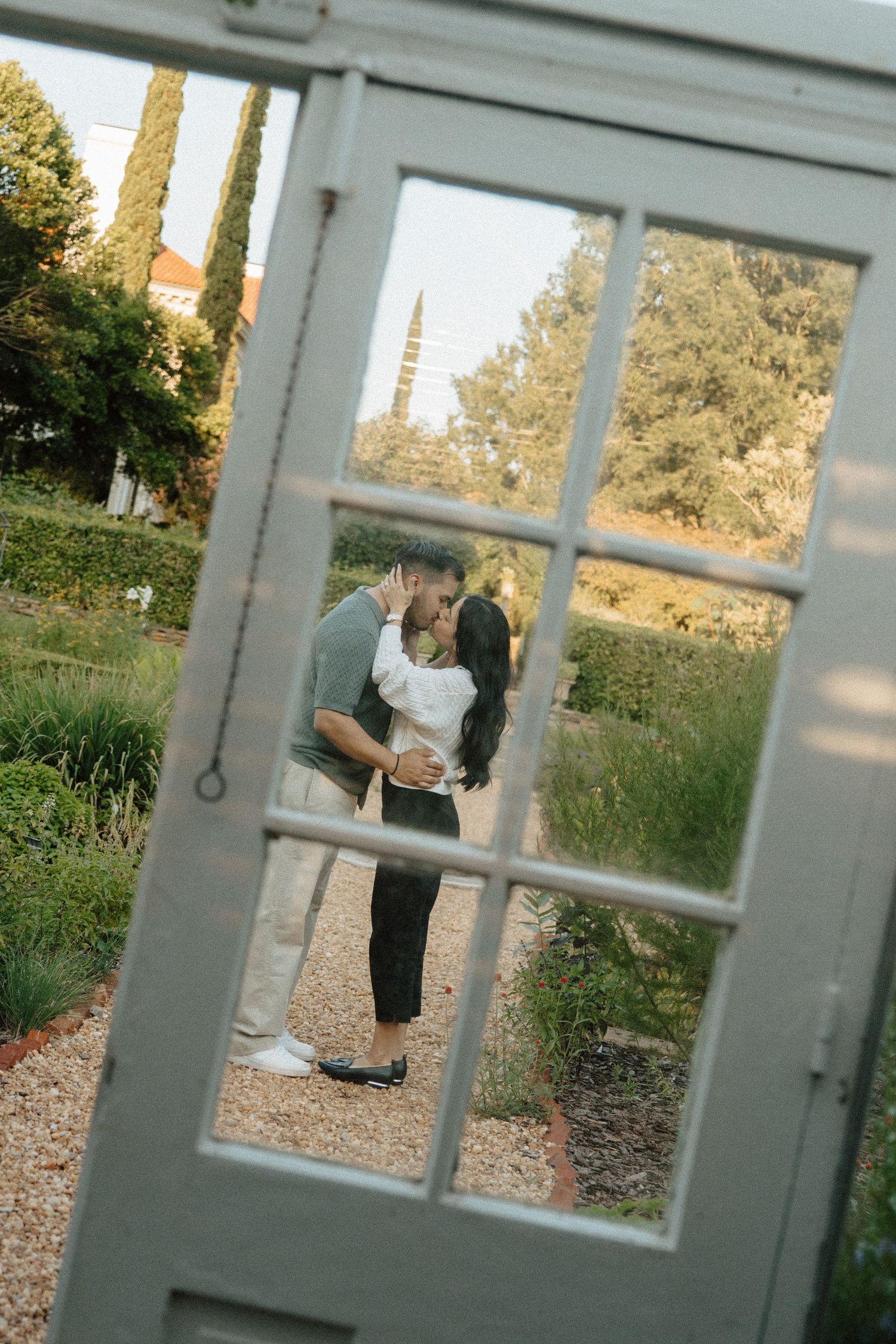 Couple sharing a romantic kiss framed through a white window in a lush garden setting.