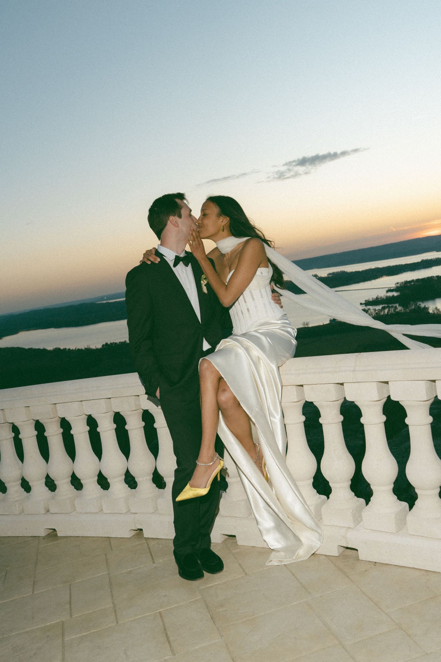 Bride and groom share a romantic kiss on a balcony at sunset, with a scenic landscape view behind them. Film Photo