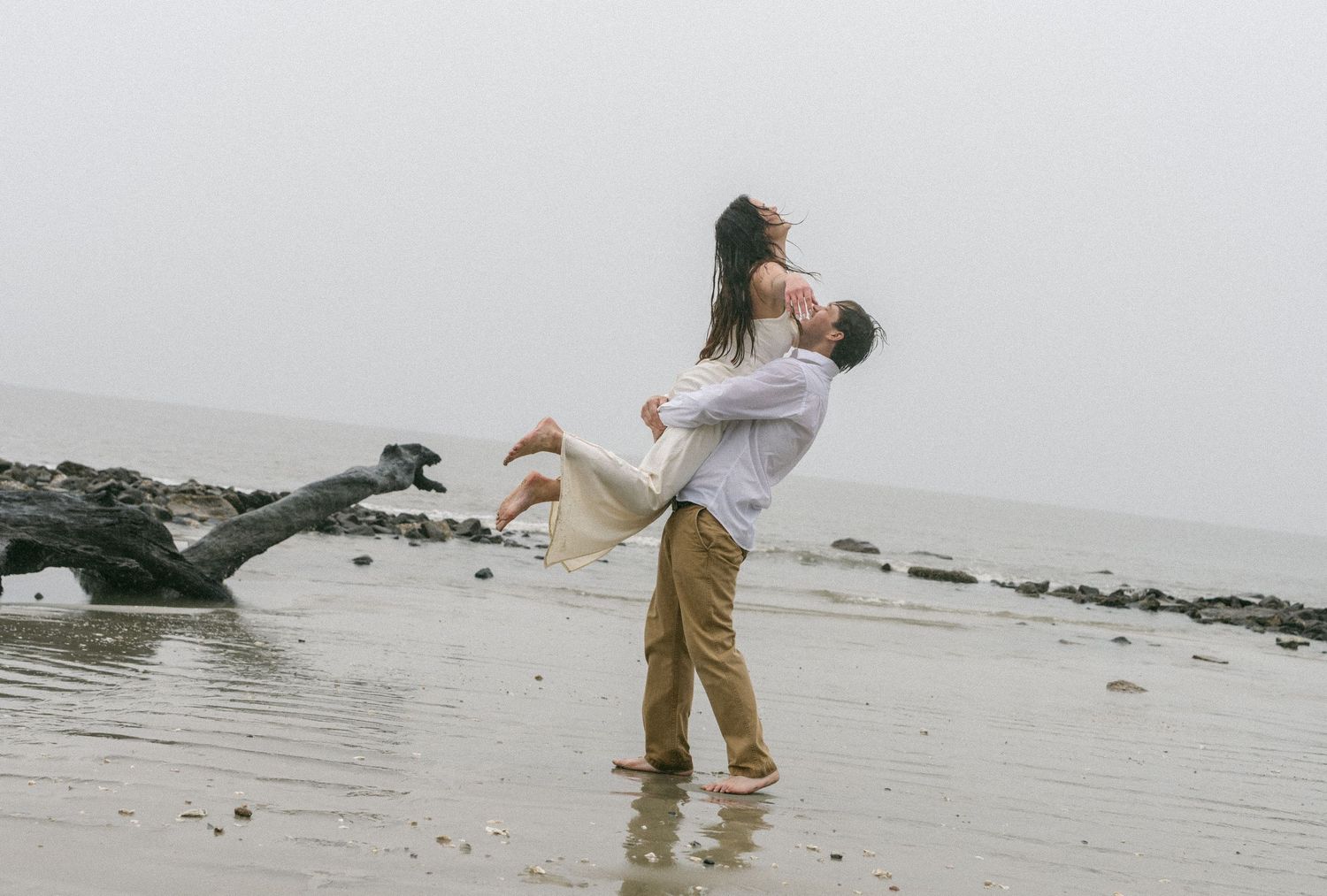 Jekyll Island Engagement. A couple shares a romantic moment on a misty beach, as the man lifts the woman in his arms near the shoreline.