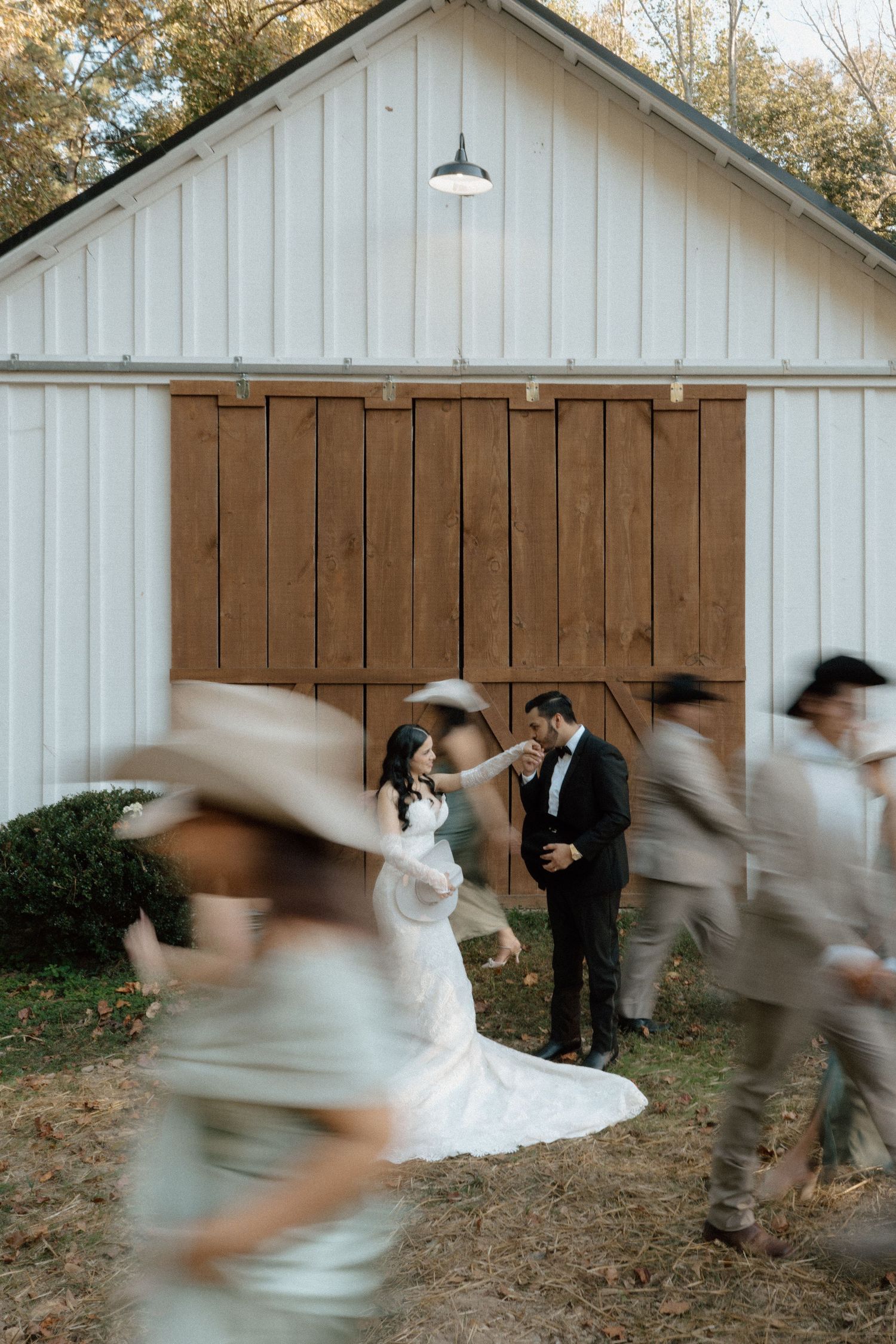 Bride and groom dance outside a white barn as blurred wedding party members move around them in a dreamy motion effect.