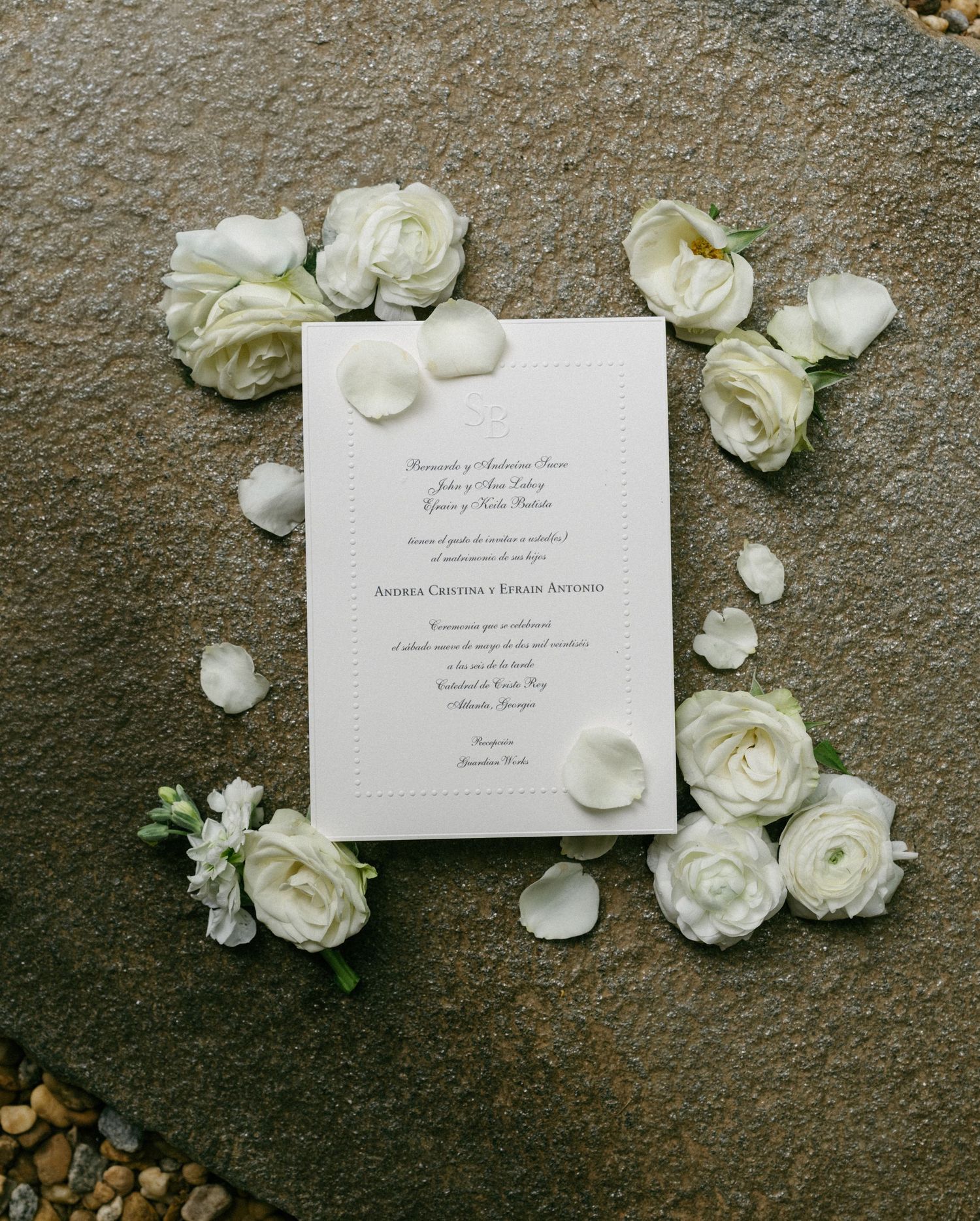 Elegant wedding invitation surrounded by white roses and petals on a gray stone surface.