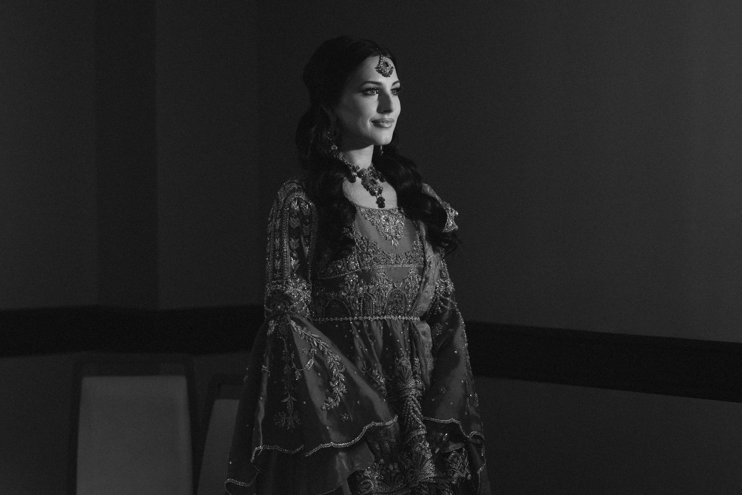 Woman in ornate traditional South Asian attire and jewelry poses gracefully in a black and white portrait.