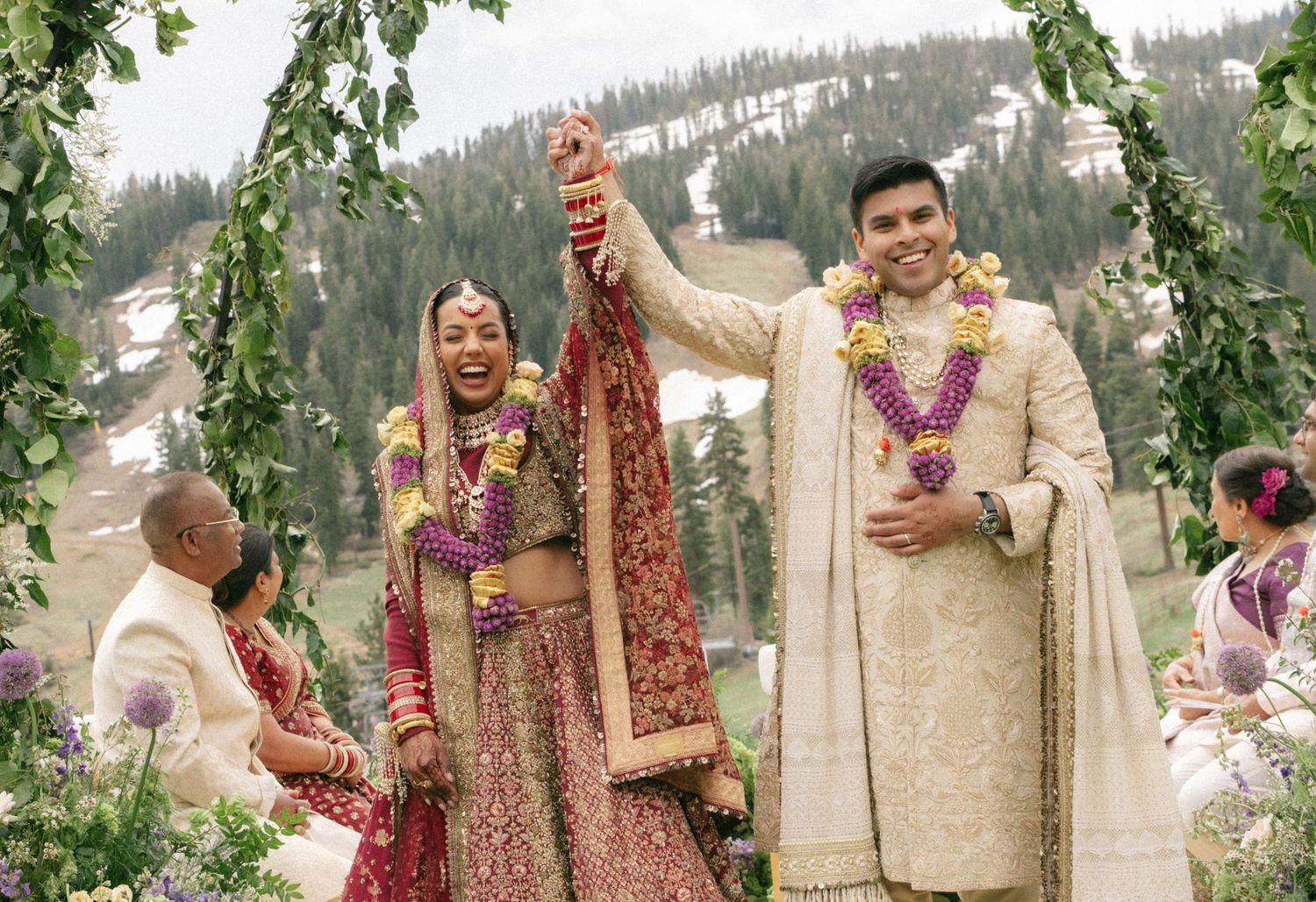 Joyful Indian bride and groom in traditional attire celebrate under floral arch at outdoor mountain wedding ceremony.