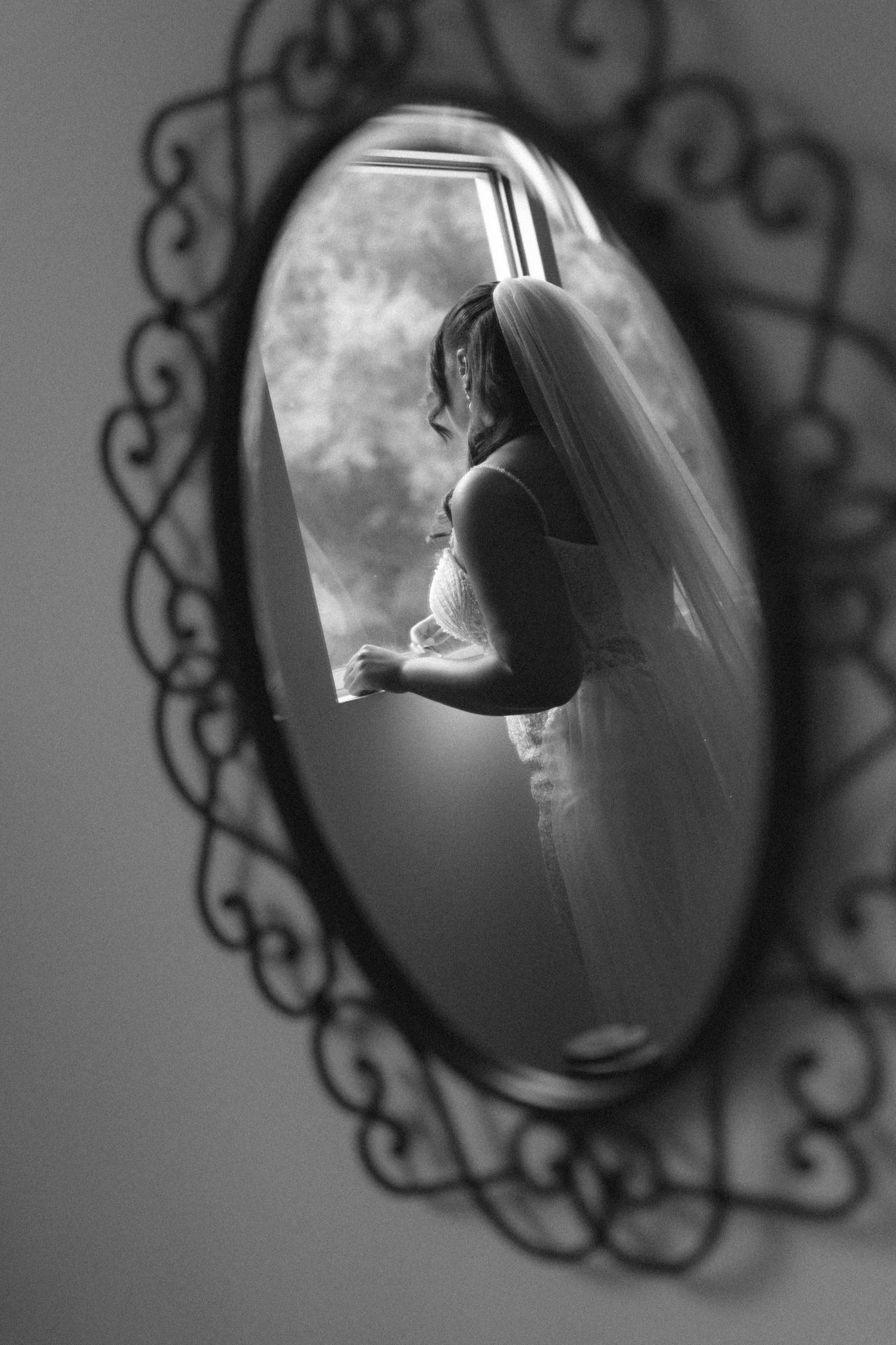 Elegant bride in wedding dress and veil reflected in ornate oval mirror in black and white photo.
