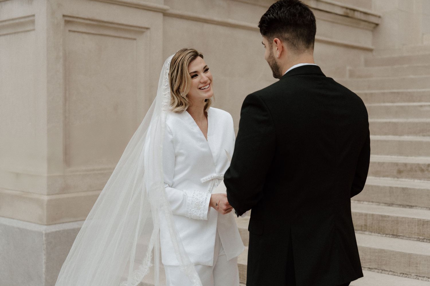 Bride in white pantsuit and veil holds hands with groom in black tuxedo on stone steps.
