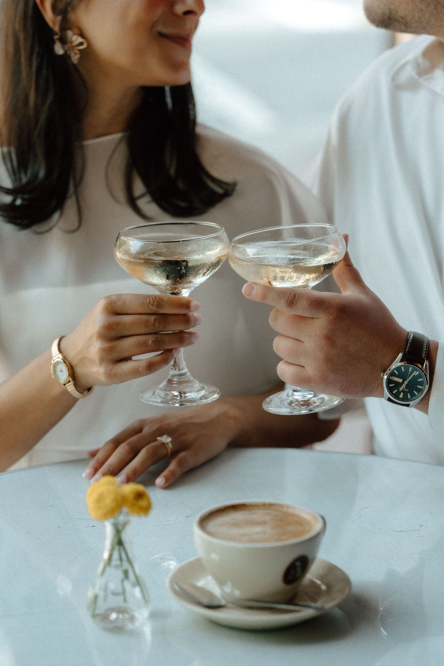 Couple clinking champagne coupe glasses at a cafe table with a latte and yellow flowers.