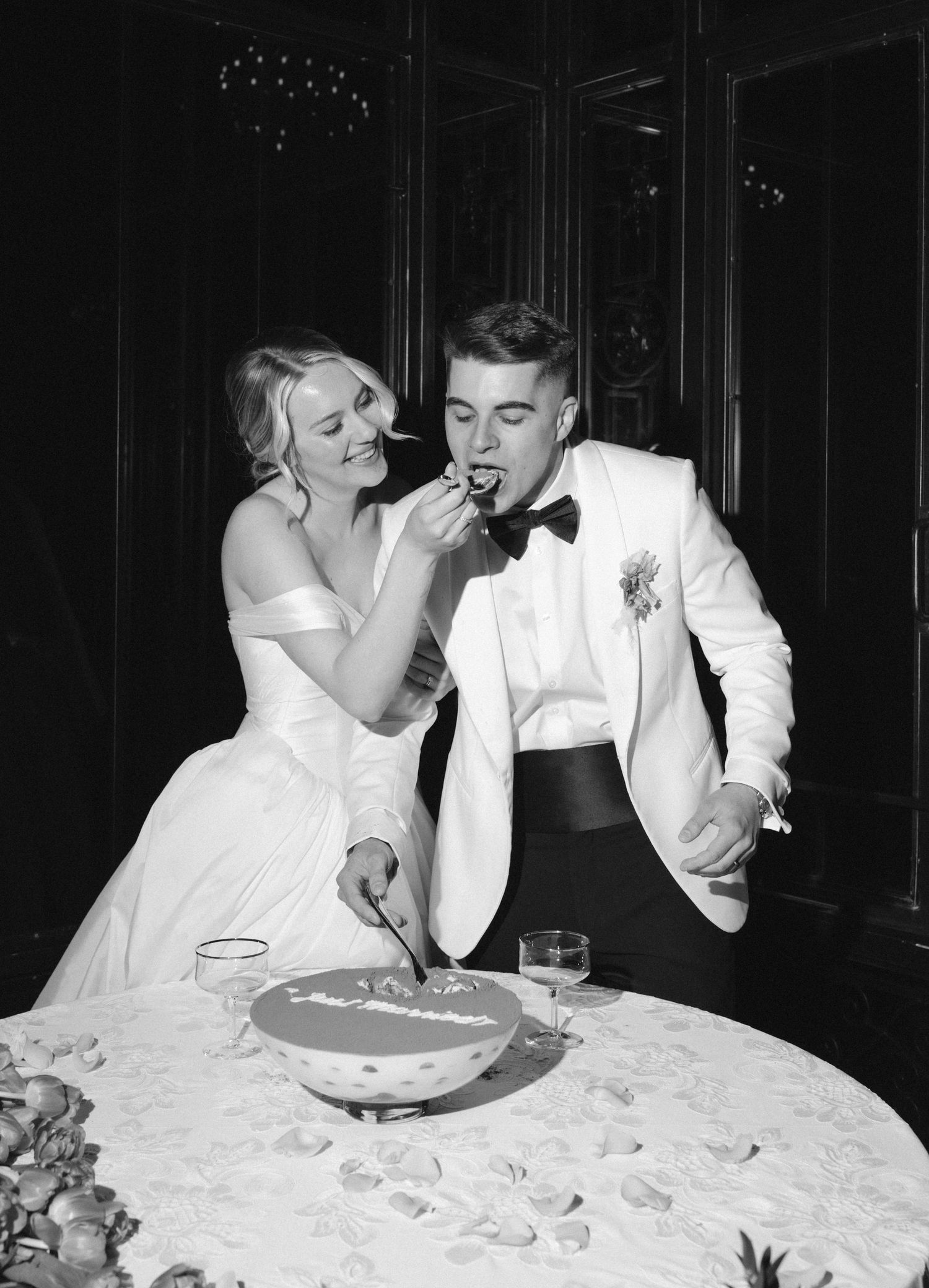 Black and white photo of bride feeding groom wedding cake at elegant reception table.