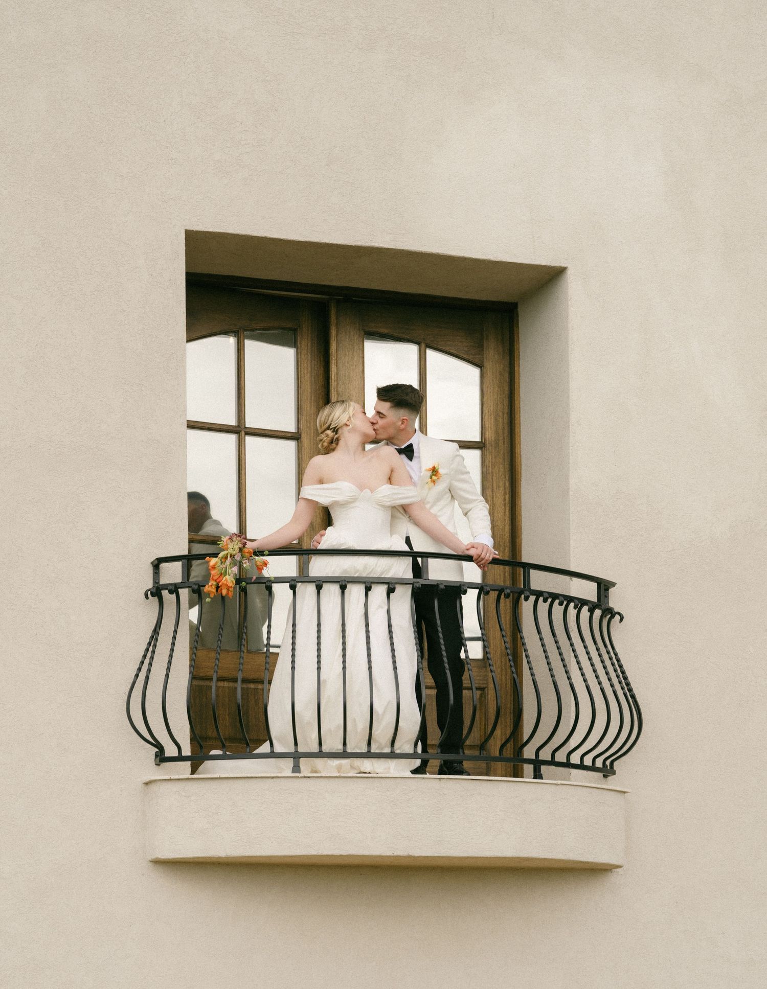 Bride and groom sharing a kiss on an elegant wrought iron balcony, bride holding a colorful bouquet.