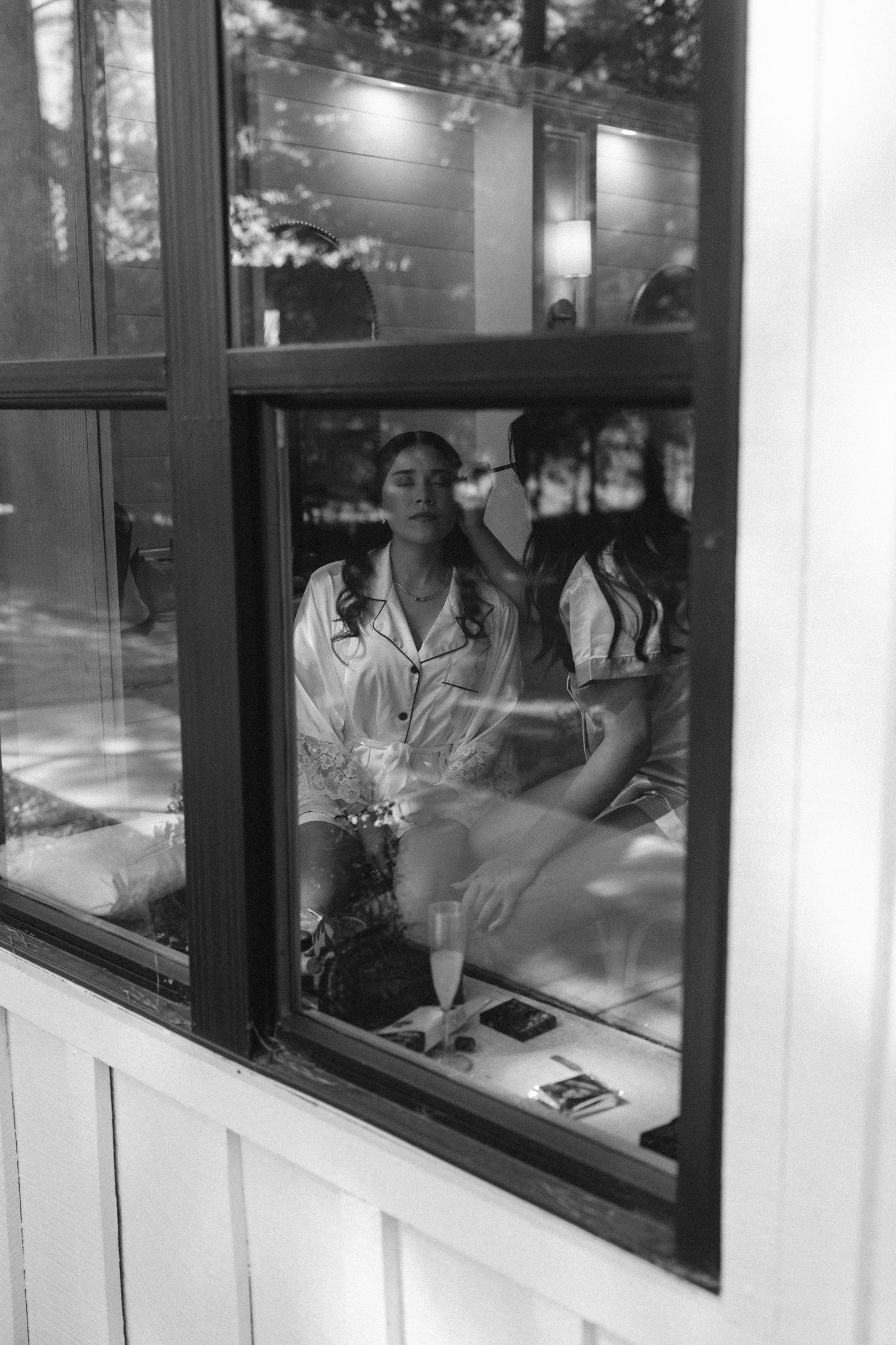 Black and white photo of two women in satin robes getting ready, viewed through a window.