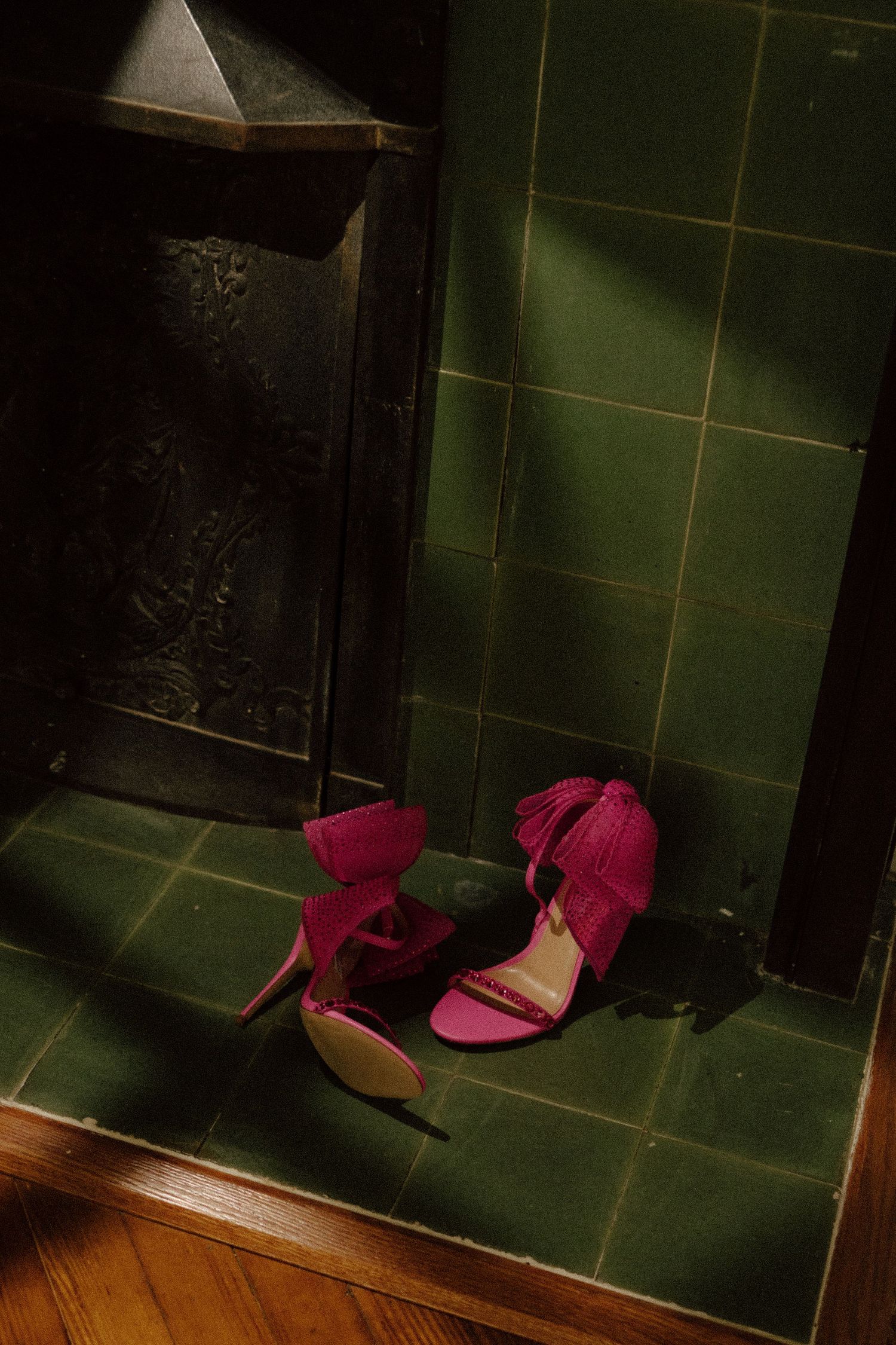 Pink fuchsia suede heeled sandals with fringe detail resting on green tiled floor near dark ornate fireplace.