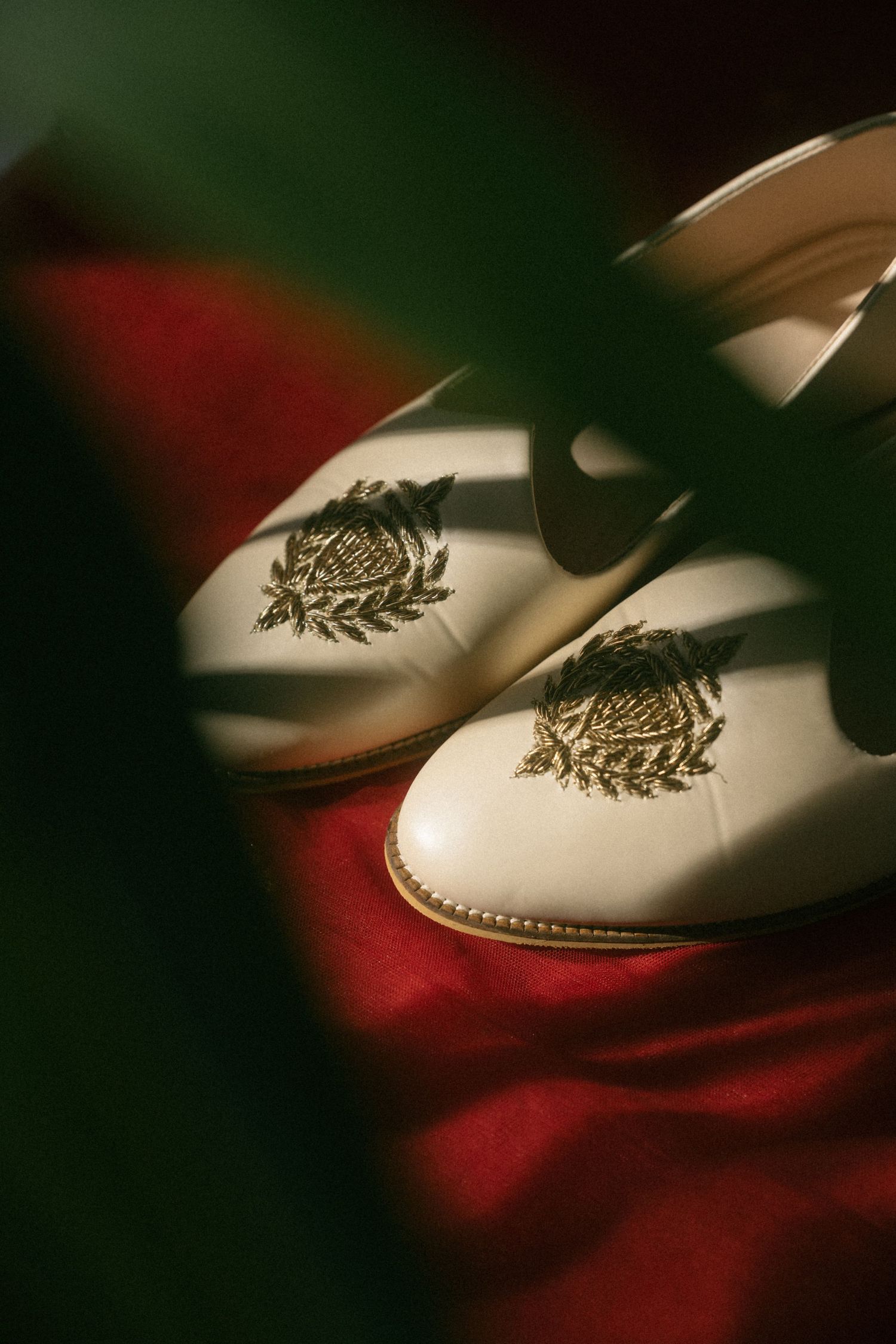 Elegant cream wedding shoes with gold embroidered floral crest resting on red silk fabric.