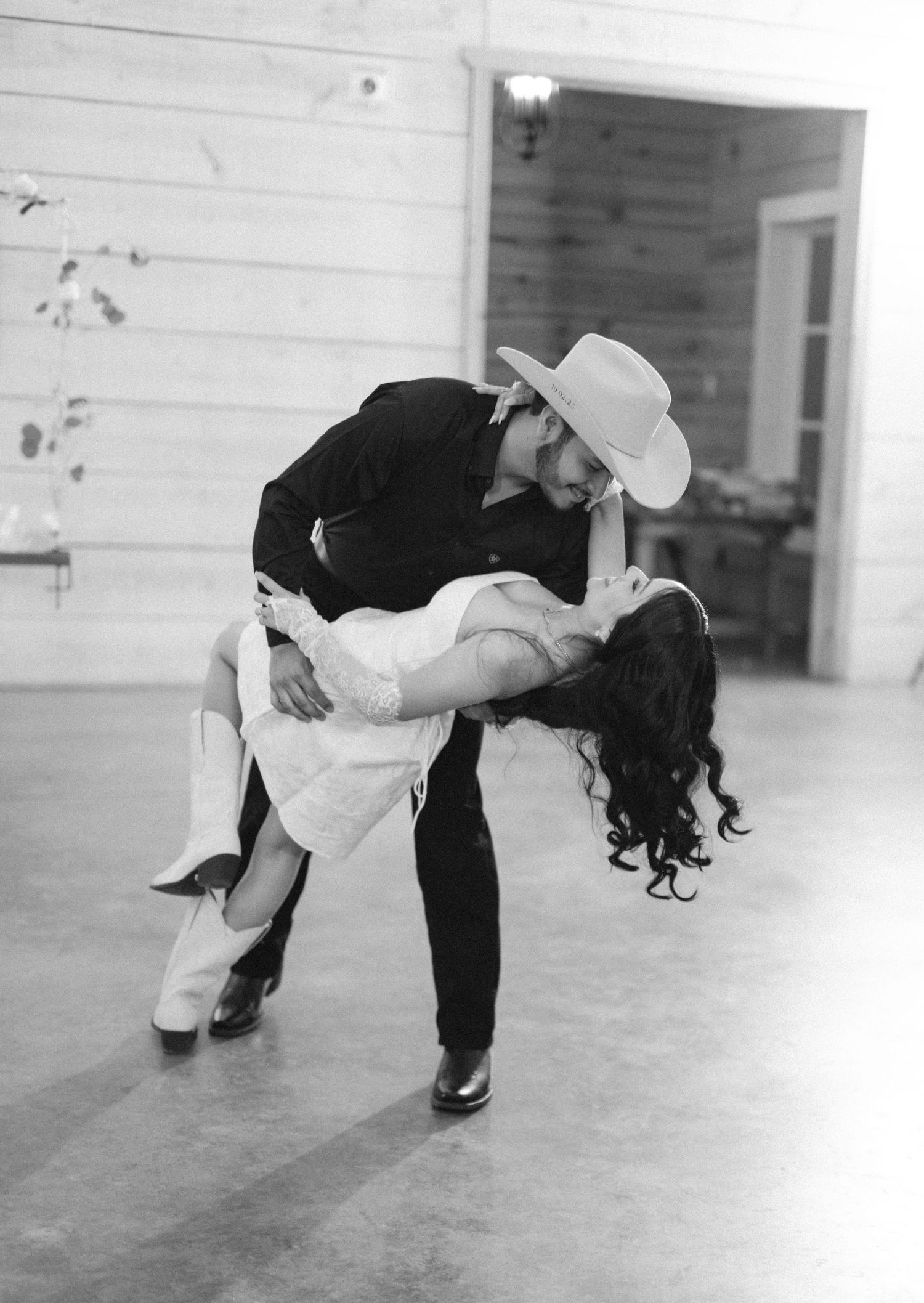 Black and white photo of a couple sharing a romantic dip dance, groom in cowboy hat and black shirt.
