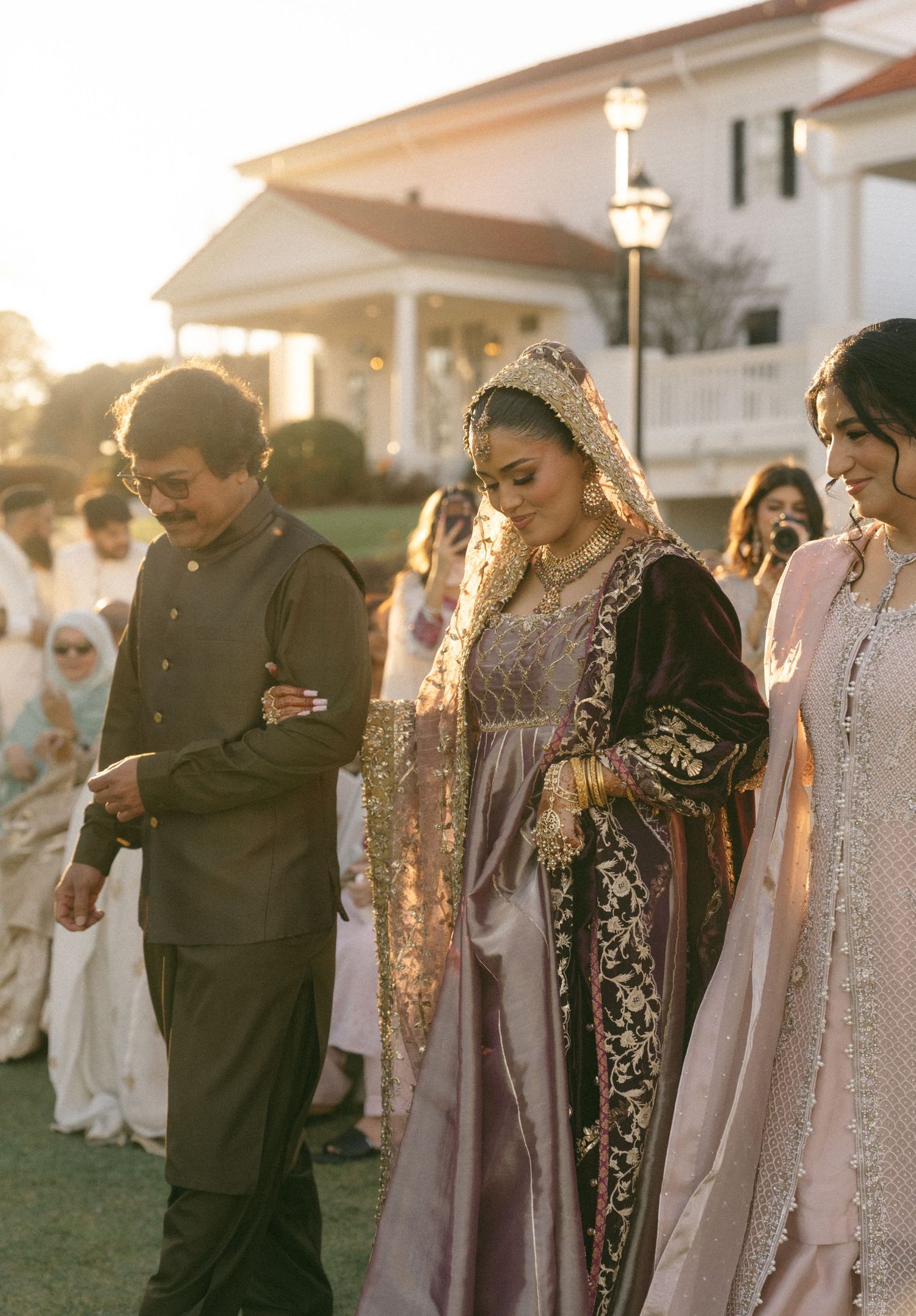 A bride in ornate Pakistani bridal dress walks with family at golden hour outside a white estate.