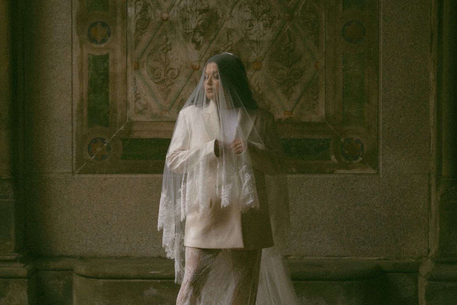 Woman in white lace veil and cream outfit standing in ornate historic interior.