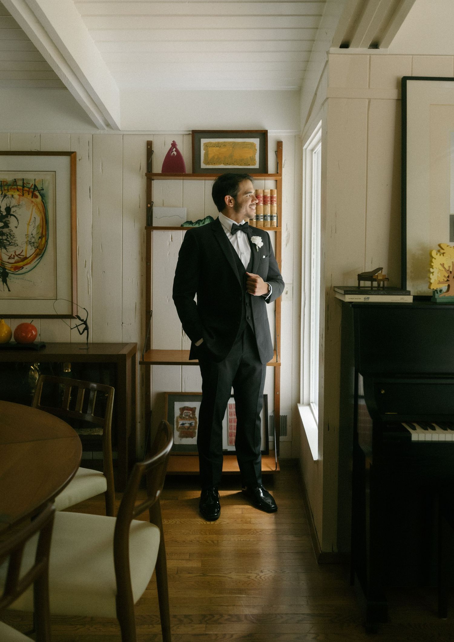 Groom in black tuxedo with boutonniere stands by window in rustic room on wedding day.