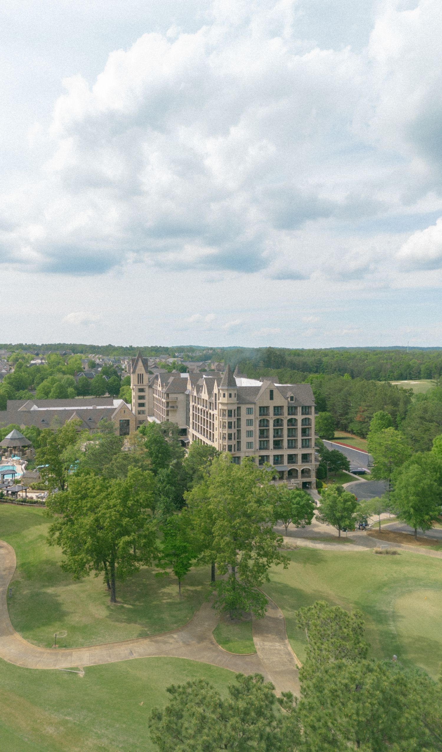 Aerial view of a grand hotel resort surrounded by lush green trees and a golf course under a partly cloudy sky.