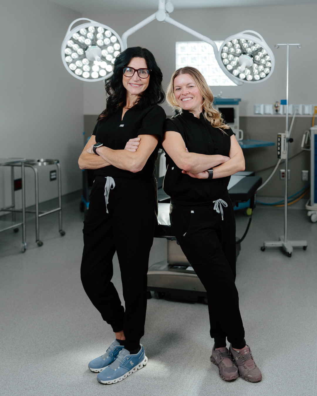 Two medical professionals in black scrubs pose confidently in a modern operating room with surgical lights overhead.