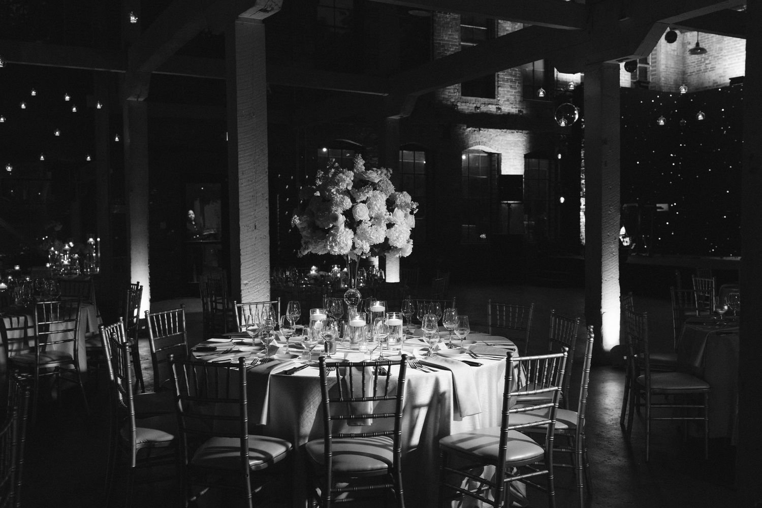 Black and white photo of a romantic wedding reception with tall floral centerpieces and string lights.