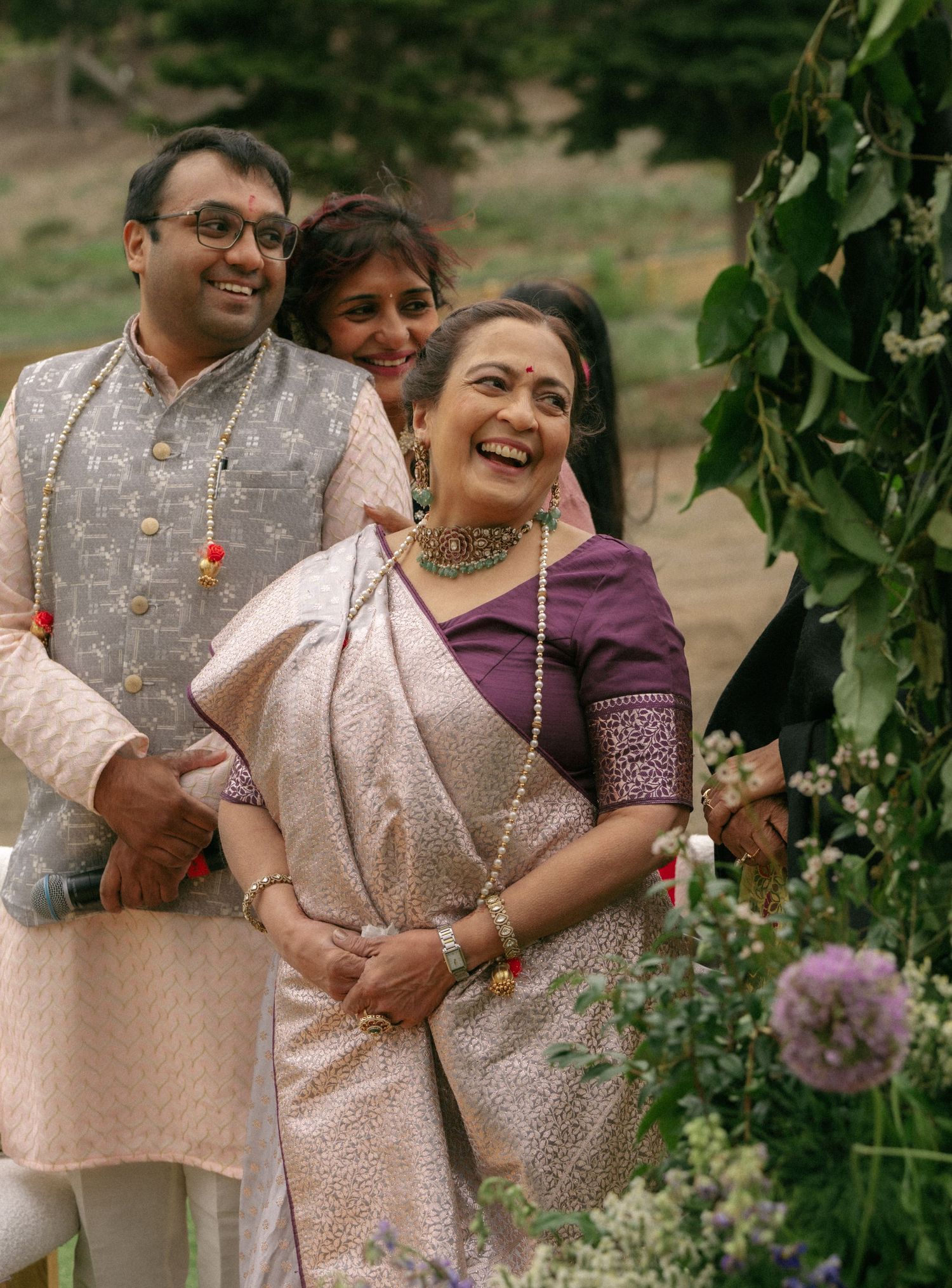 Smiling Indian woman in purple blouse and white saree enjoys outdoor wedding celebration with family.