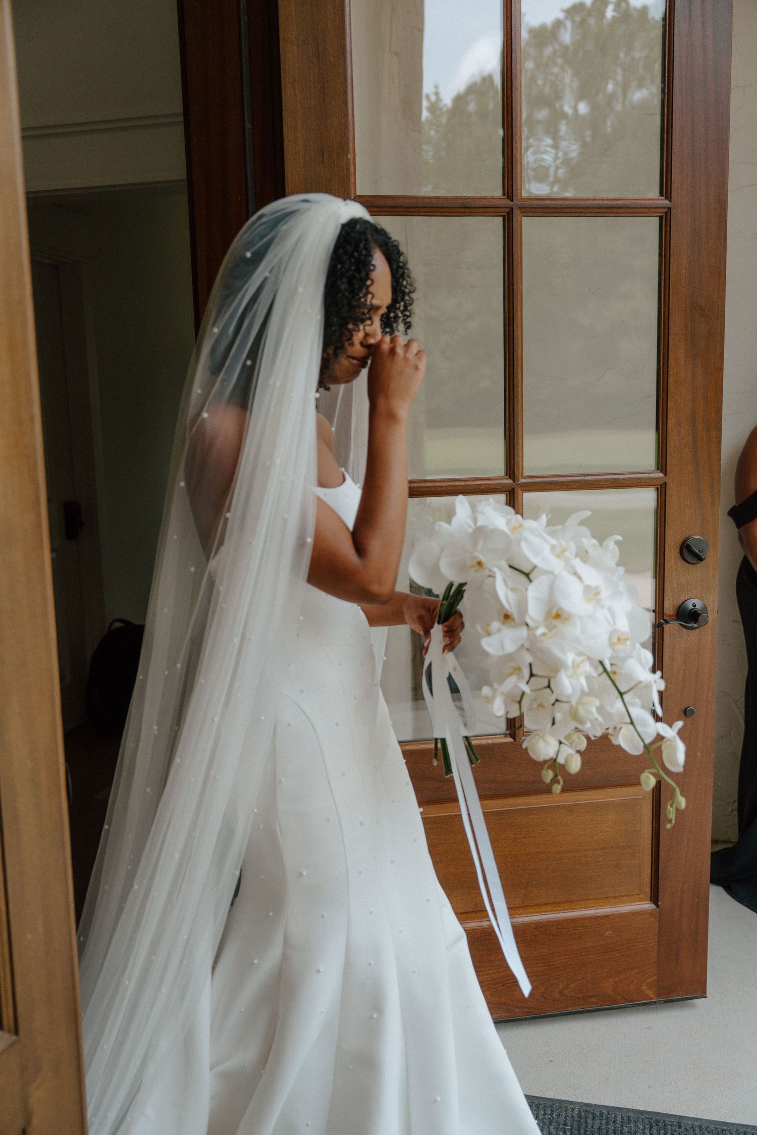 Bride in white gown and veil holding white orchid bouquet near wooden door.