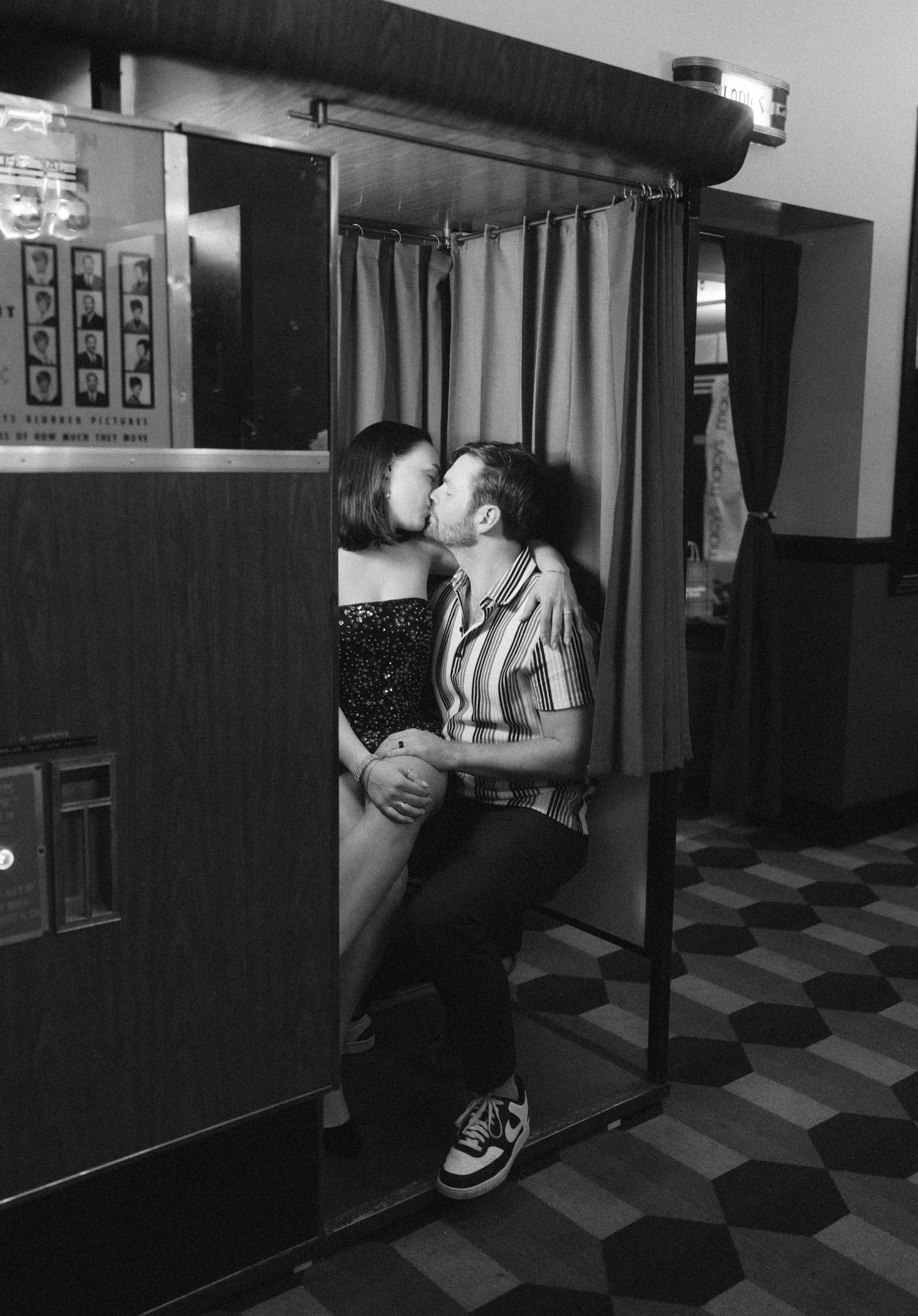 Couple kissing in a vintage photo booth, black and white romantic moment captured inside curtained enclosure.