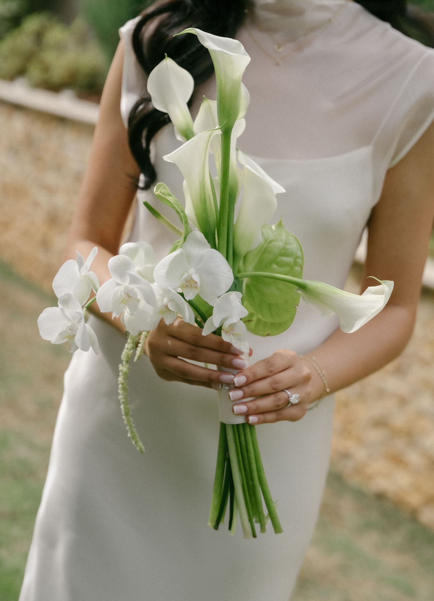Bride in white satin dress holding elegant bouquet of white calla lilies and orchids with green anthurium leaves.