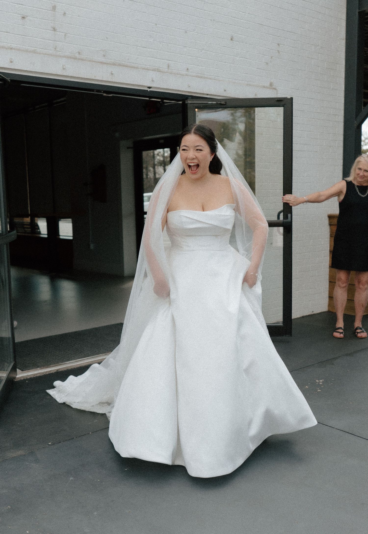 Excited bride in strapless white gown and veil laughing joyfully at venue entrance on wedding day.