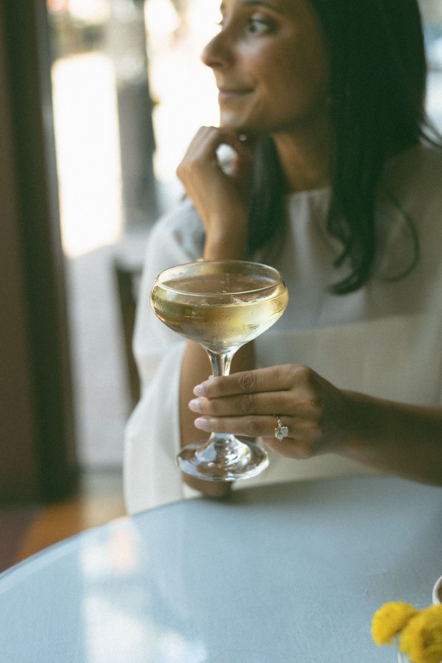 Woman holding a coupe glass of white wine at a table, wearing an engagement ring, smiling softly.