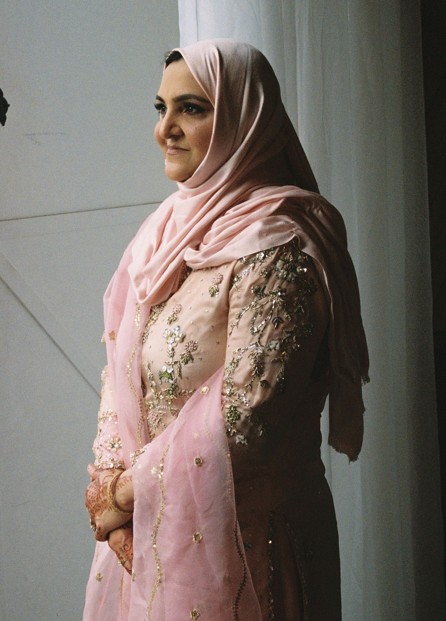 Woman in pink hijab and embroidered South Asian dress with mehndi, standing gracefully by a white column.
