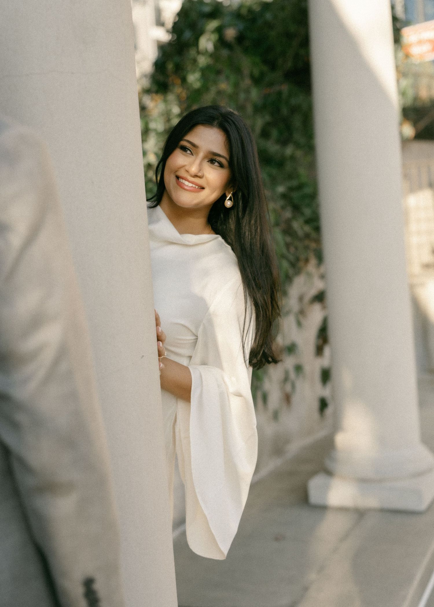 Smiling young woman in white outfit peeking around a column outdoors.