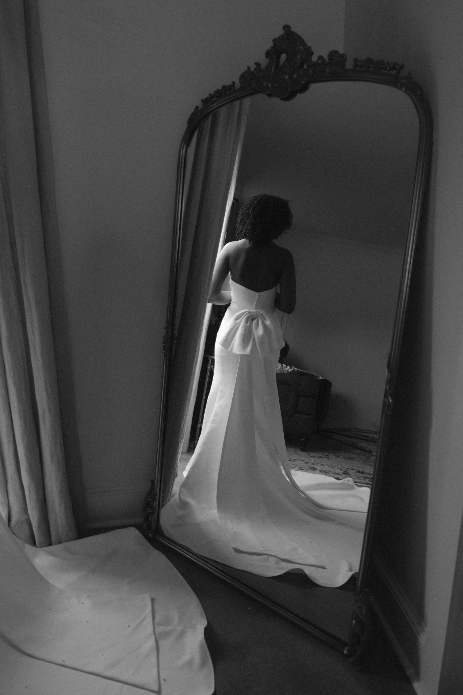 Black and white photo of bride in elegant wedding gown with train reflected in ornate vintage floor mirror.