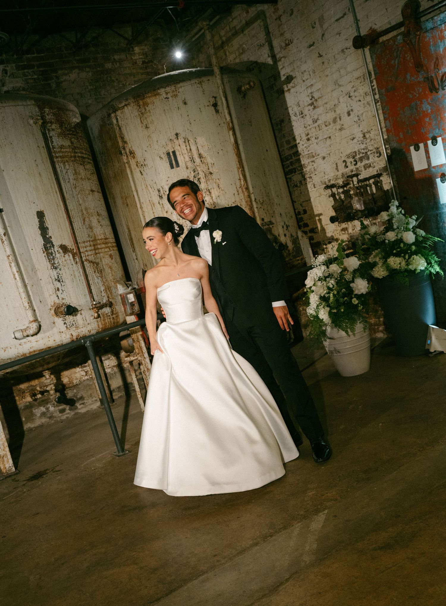 A bride in a strapless white gown and groom in a black tuxedo walk together in a rustic industrial wedding venue.