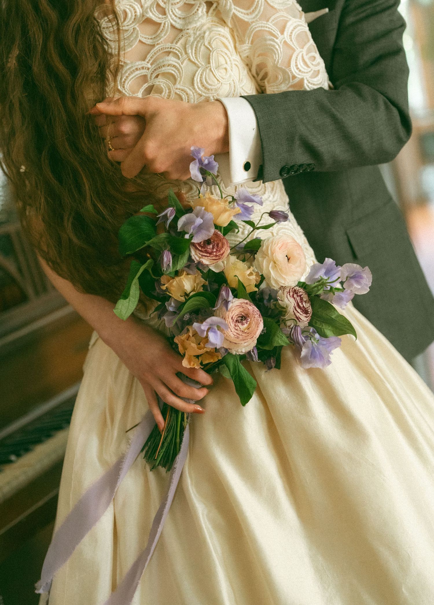 Bride holding a romantic bouquet of purple, pink, and yellow flowers with greenery, wearing an ivory gown beside a groom.