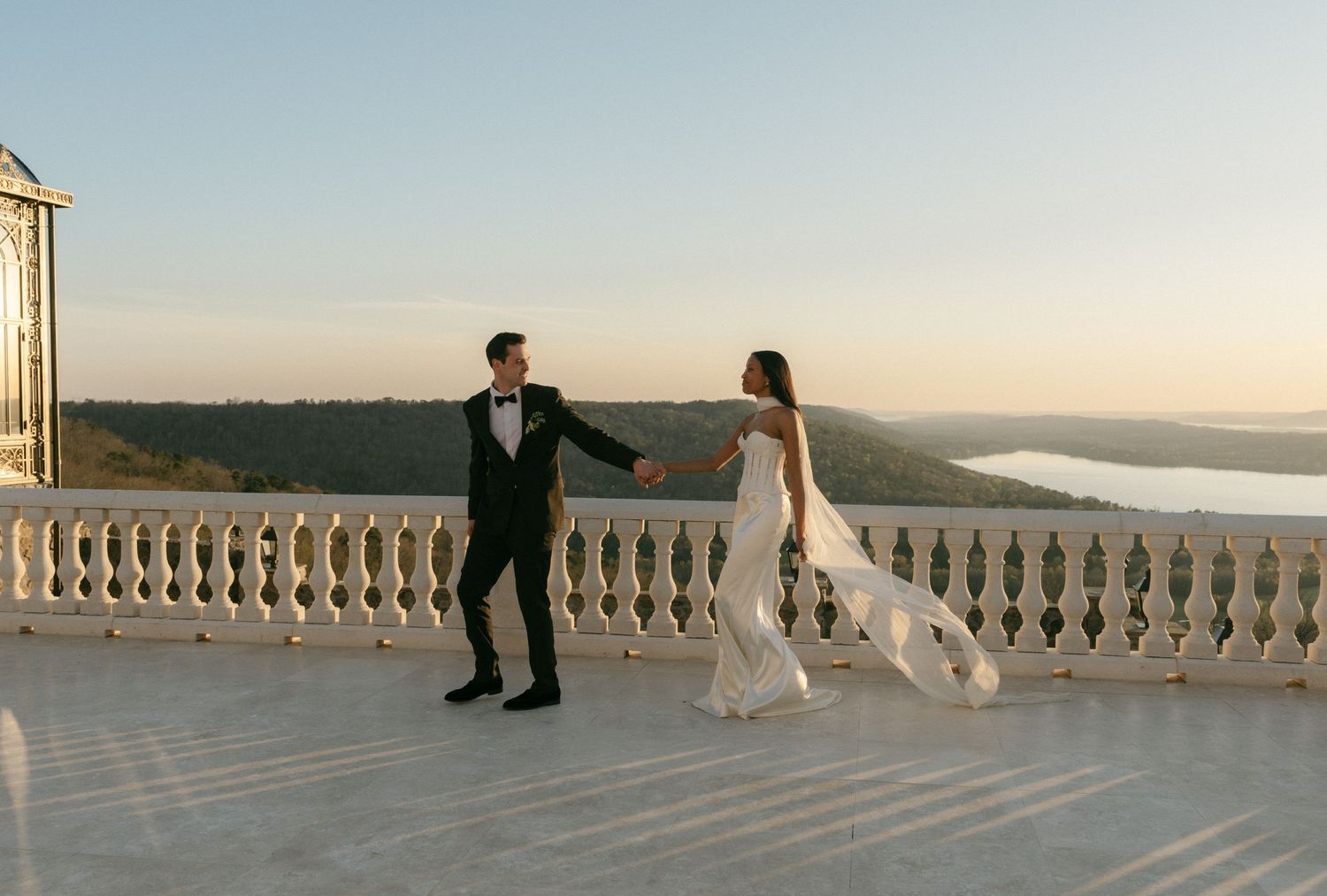 Bride and groom holding hands on terrace at sunset with scenic river valley backdrop.