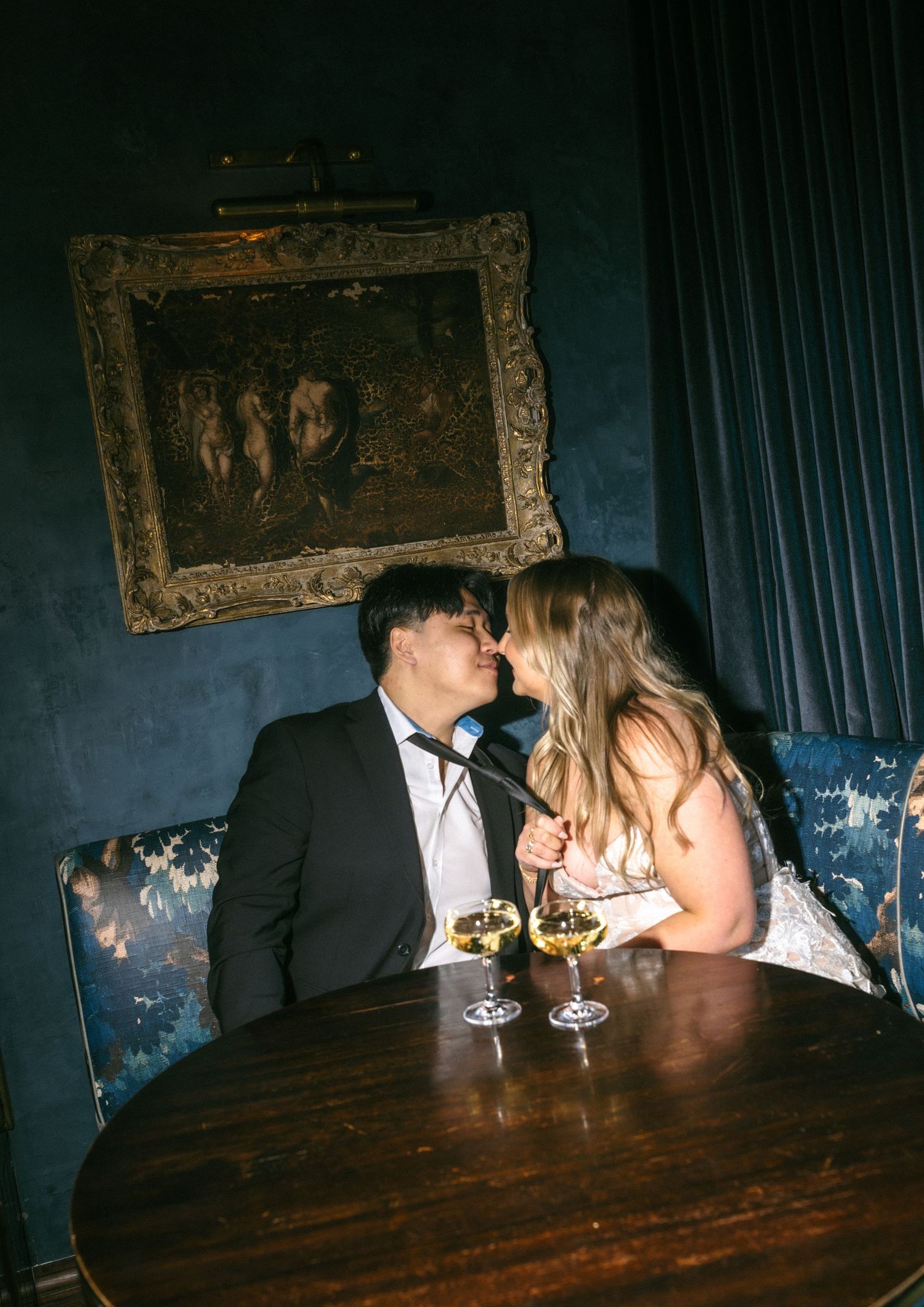 Couple sharing a romantic kiss over white wine glasses at a dark blue velvet booth in an elegant upscale bar.