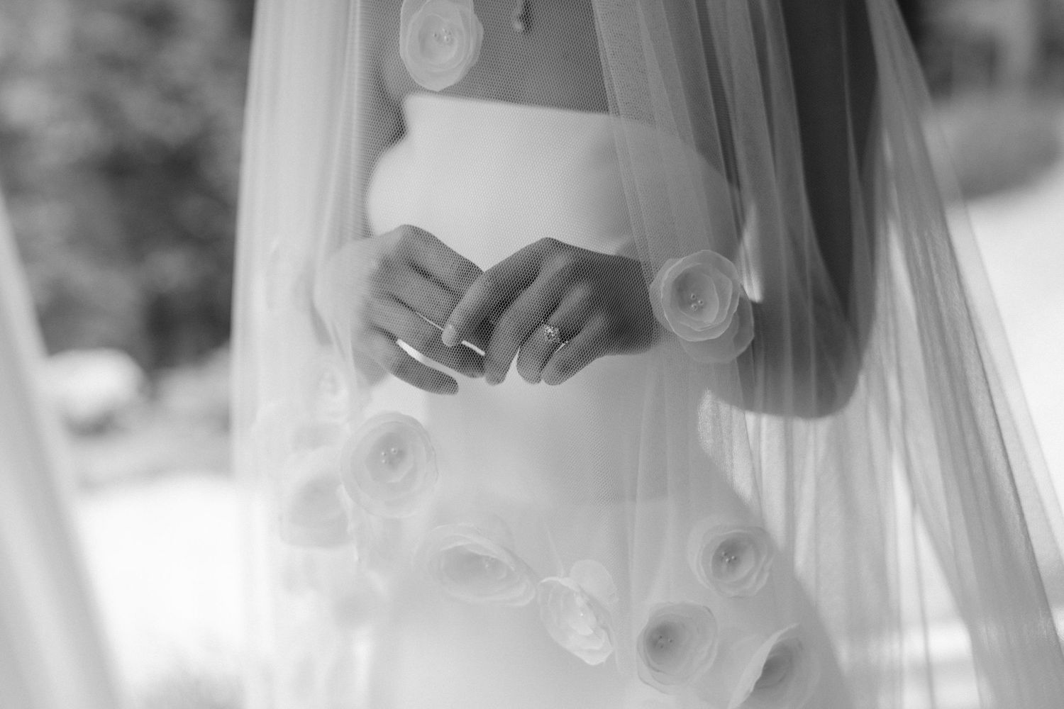 Black and white photo of a bride in a floral wedding gown with veil, hands clasped showing wedding ring.