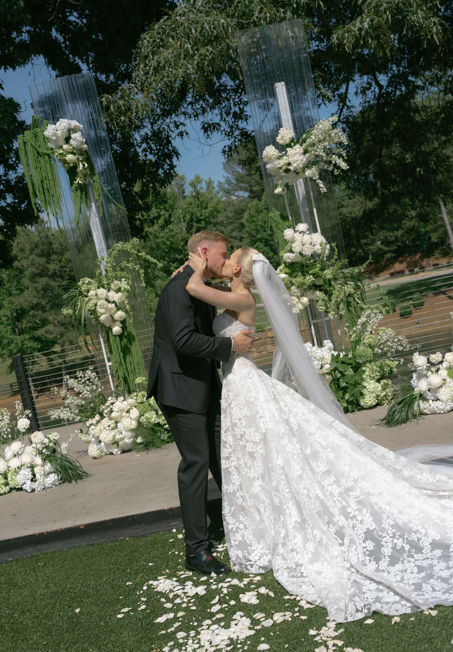 Bride and groom share first kiss at outdoor wedding ceremony surrounded by lush white floral arrangements and greenery.