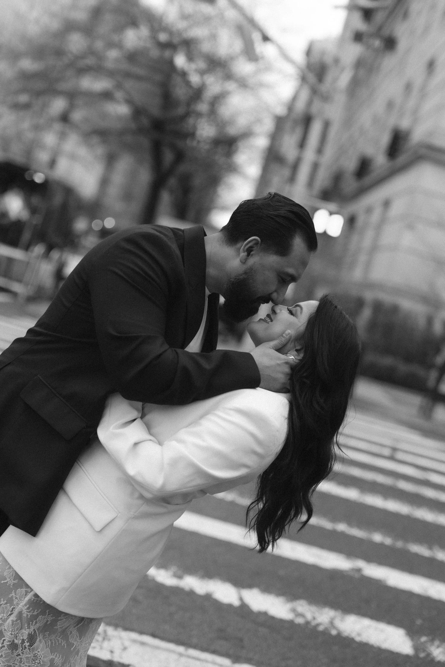 Black and white photo of a couple sharing a romantic kiss on a city street, man in suit dipping woman in white coat.