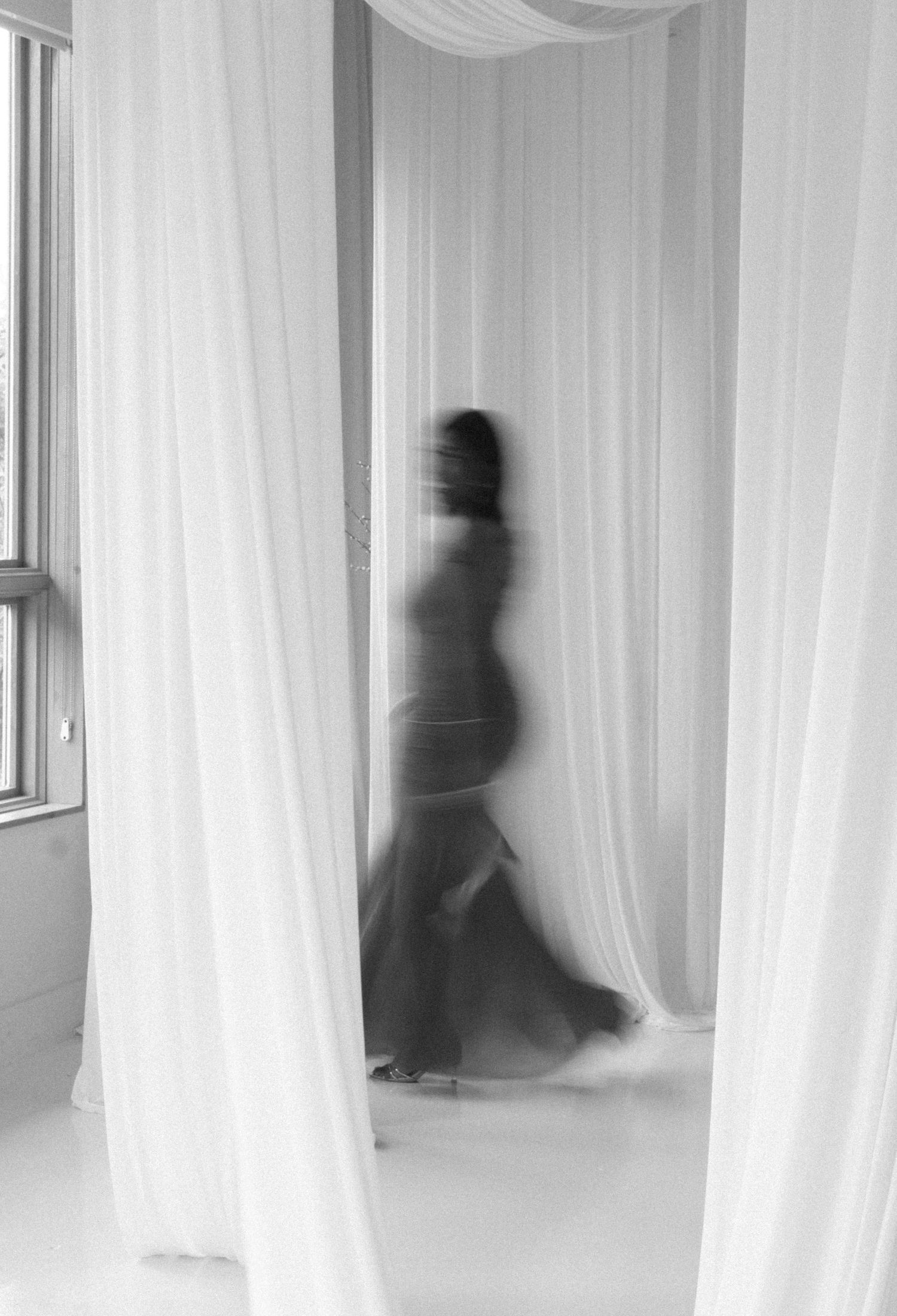 Blurred black and white photo of a woman in a flowing skirt moving gracefully between sheer white curtains.