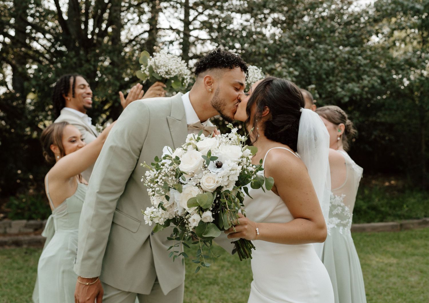 Bride and groom share a kiss surrounded by bridesmaids in sage green dresses at an outdoor wedding ceremony.