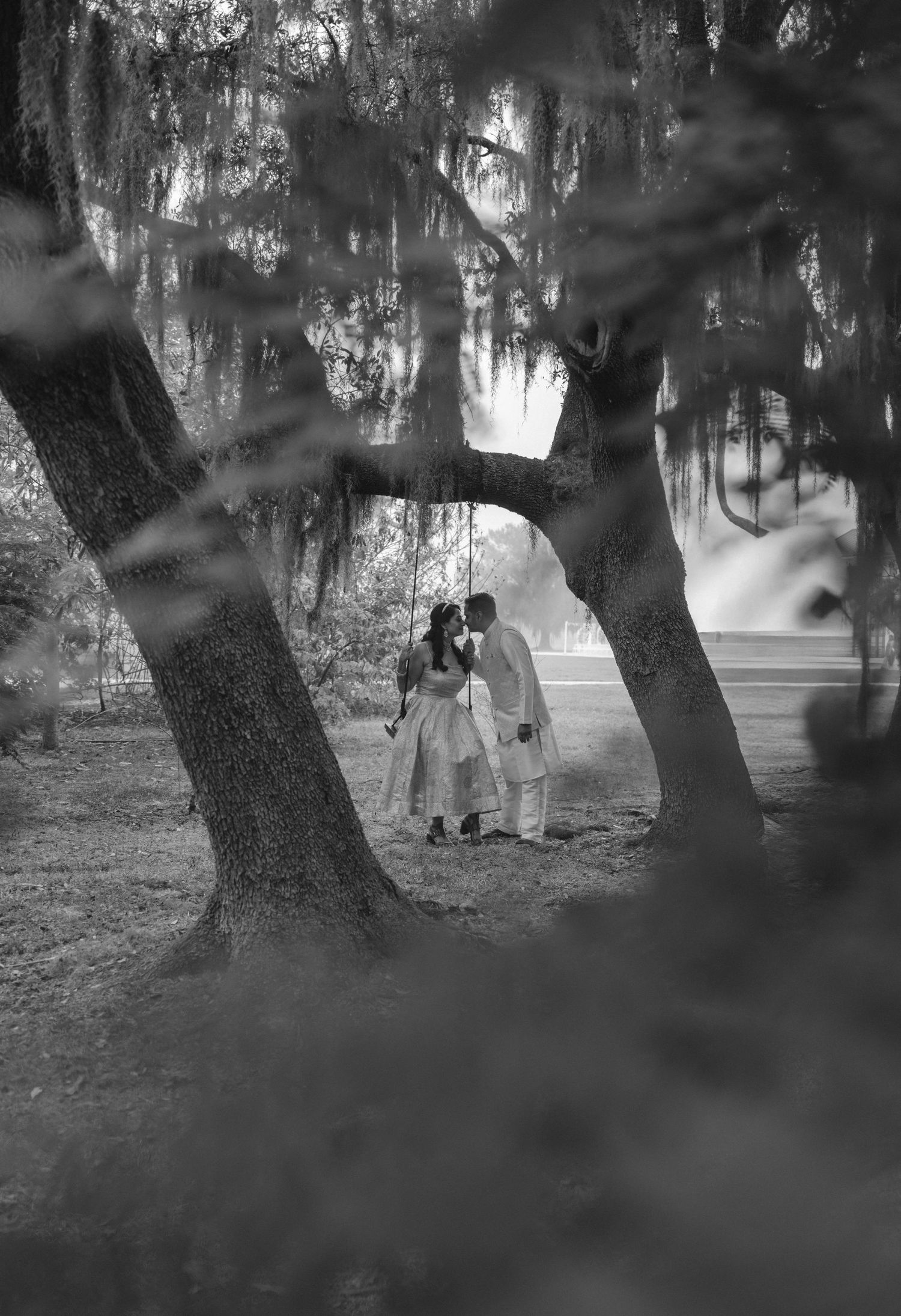 Couple sharing a romantic kiss on a swing beneath Spanish moss-draped trees in a dreamy black and white wedding photo.