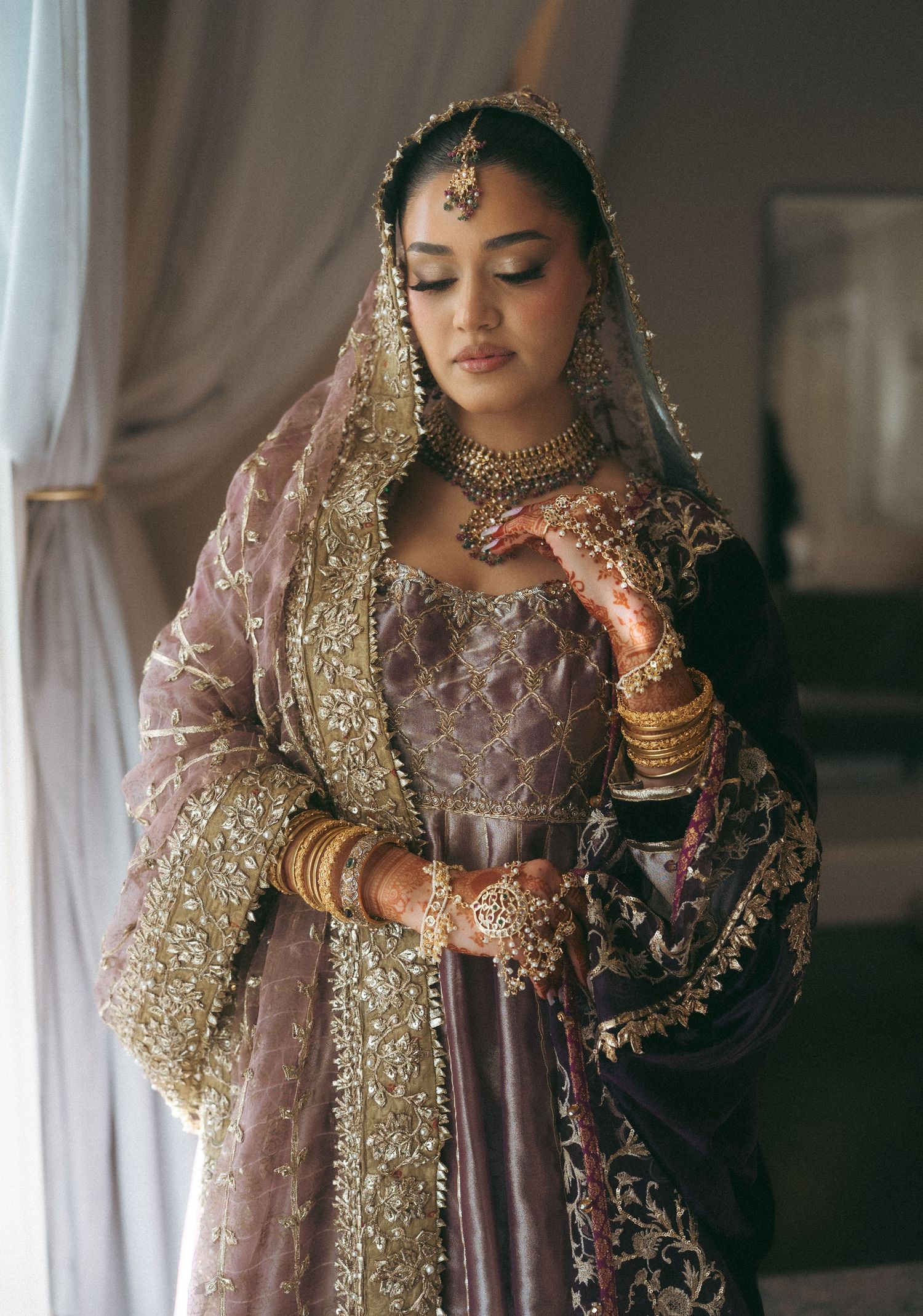 A bride in an embroidered mauve lehenga with gold dupatta, bangles, and traditional jewelry poses gracefully indoors.