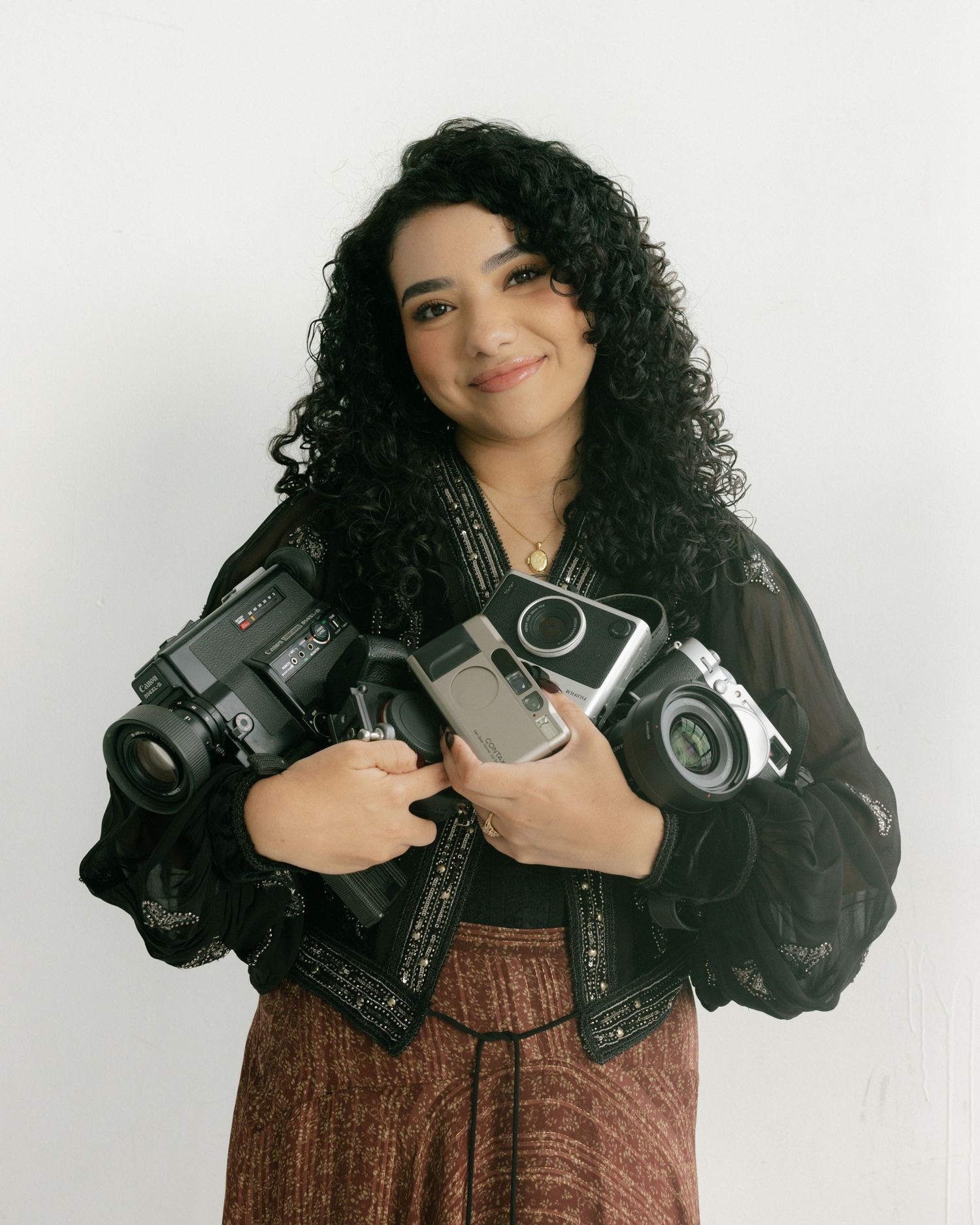 A young woman with curly dark hair holds multiple vintage cameras while smiling, wearing a black lace jacket and brown skirt.