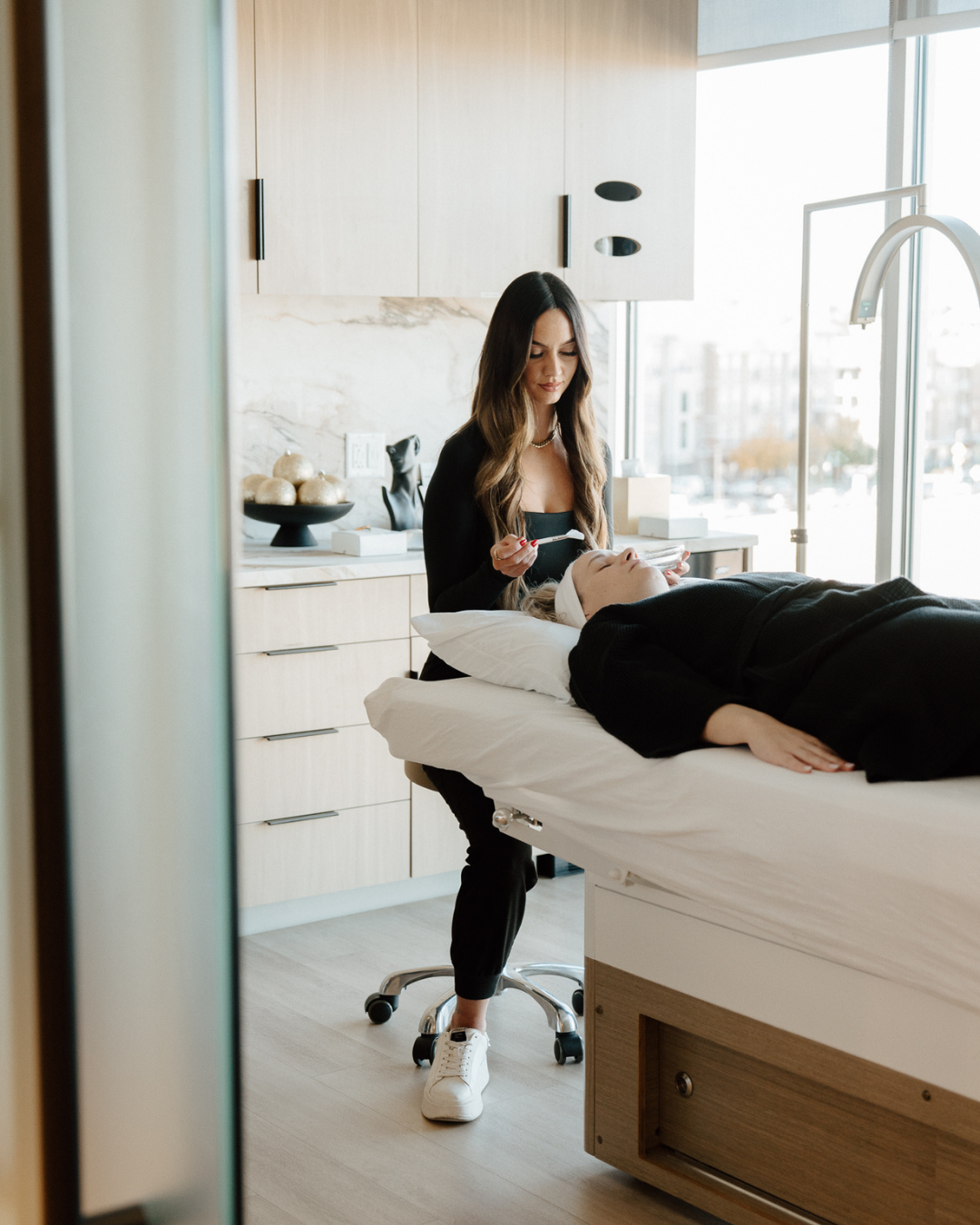 Esthetician performing a facial treatment on a client lying on a spa bed in a modern, bright treatment room.