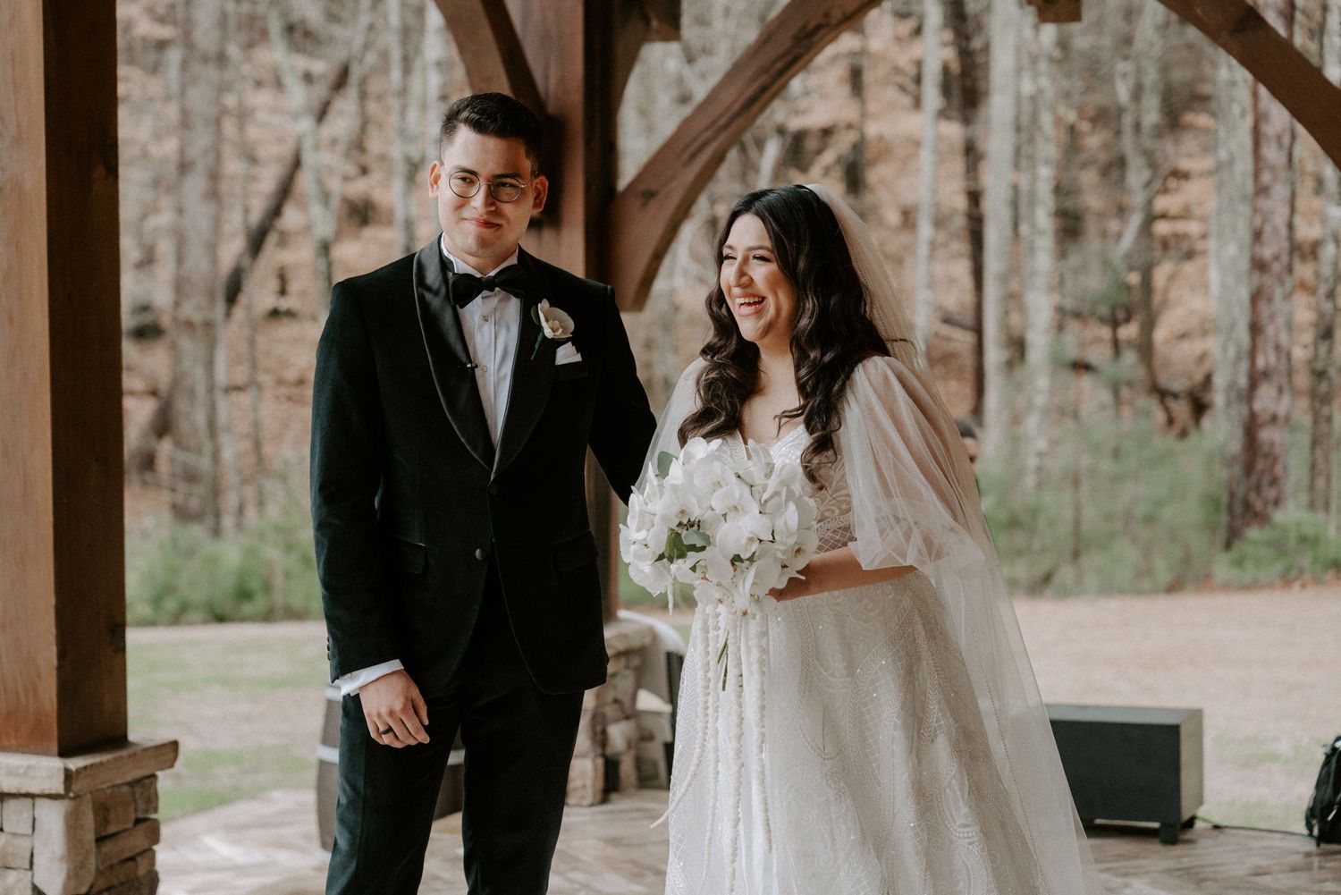 A smiling bride in a white gown and groom in a black tuxedo stand together under a wooden pavilion outdoors.