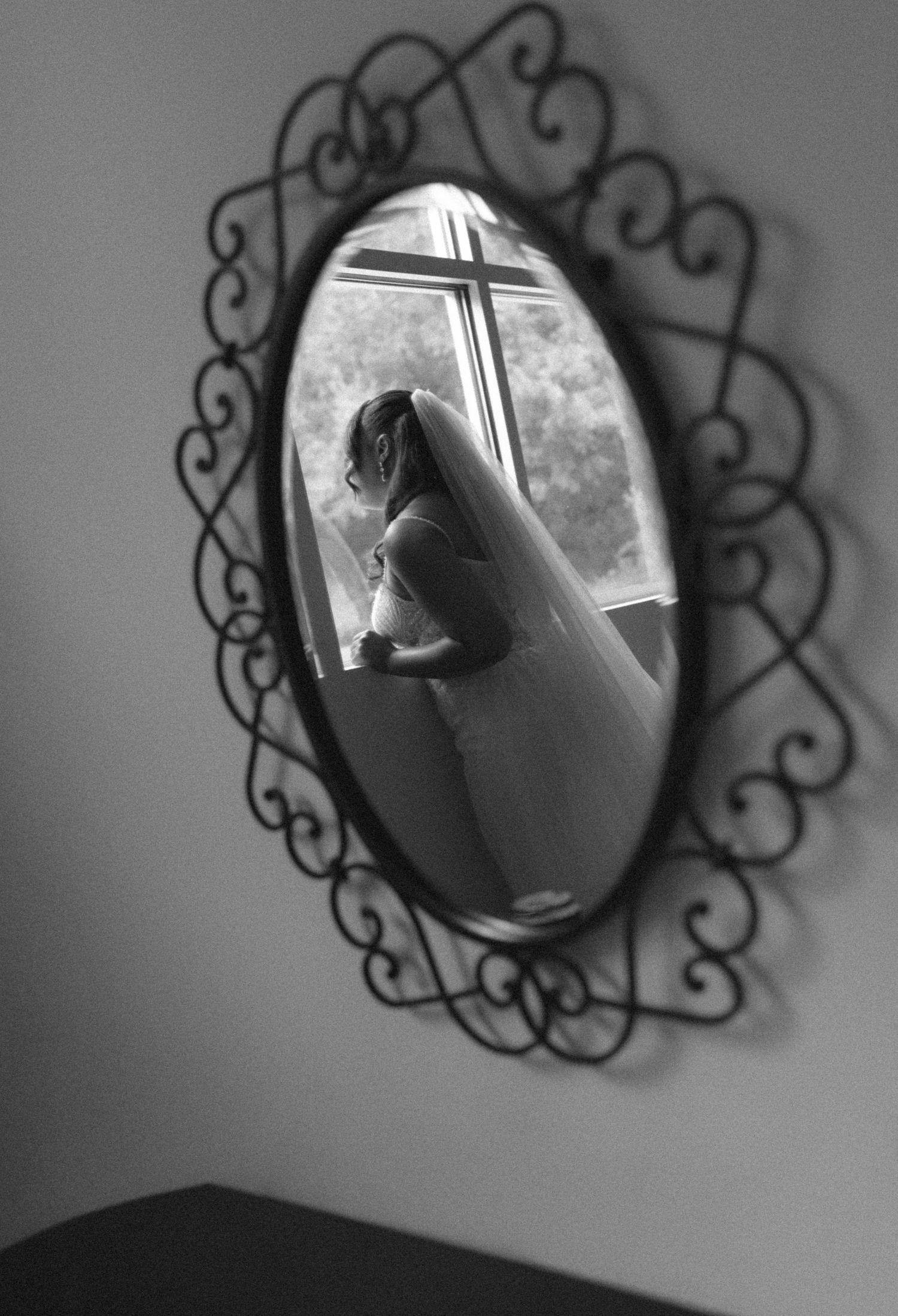 Black and white mirror reflection of a bride in wedding veil standing by a window, framed by ornate wrought iron mirror.