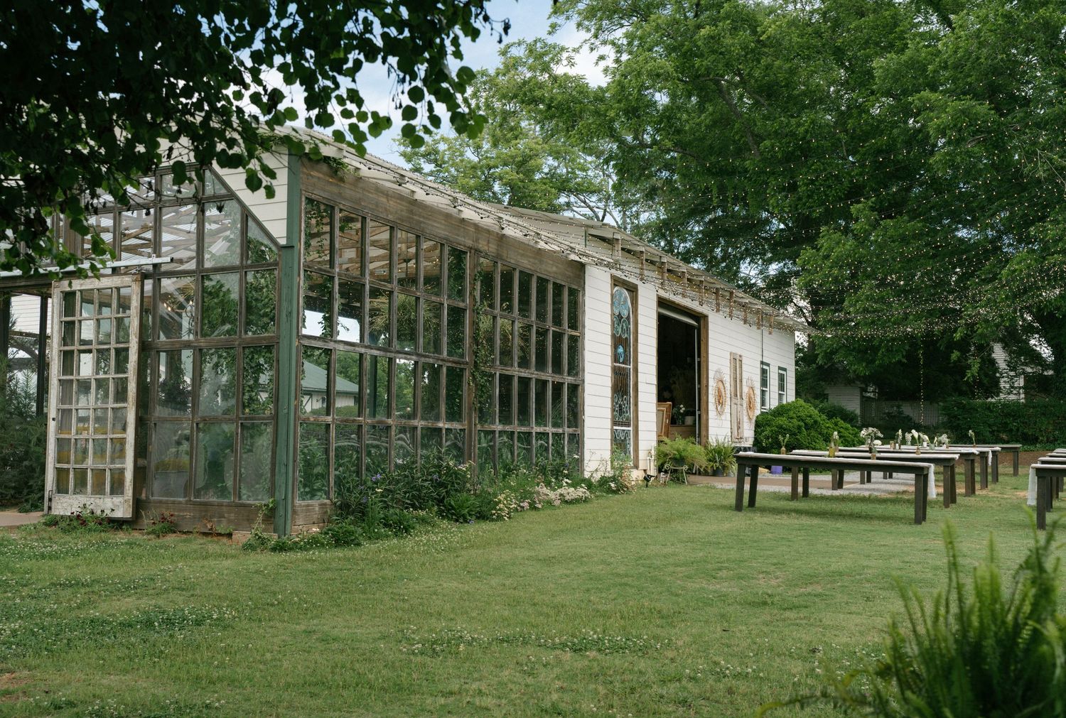 Rustic greenhouse with glass panel walls surrounded by lush green trees and manicured lawn with wooden benches.