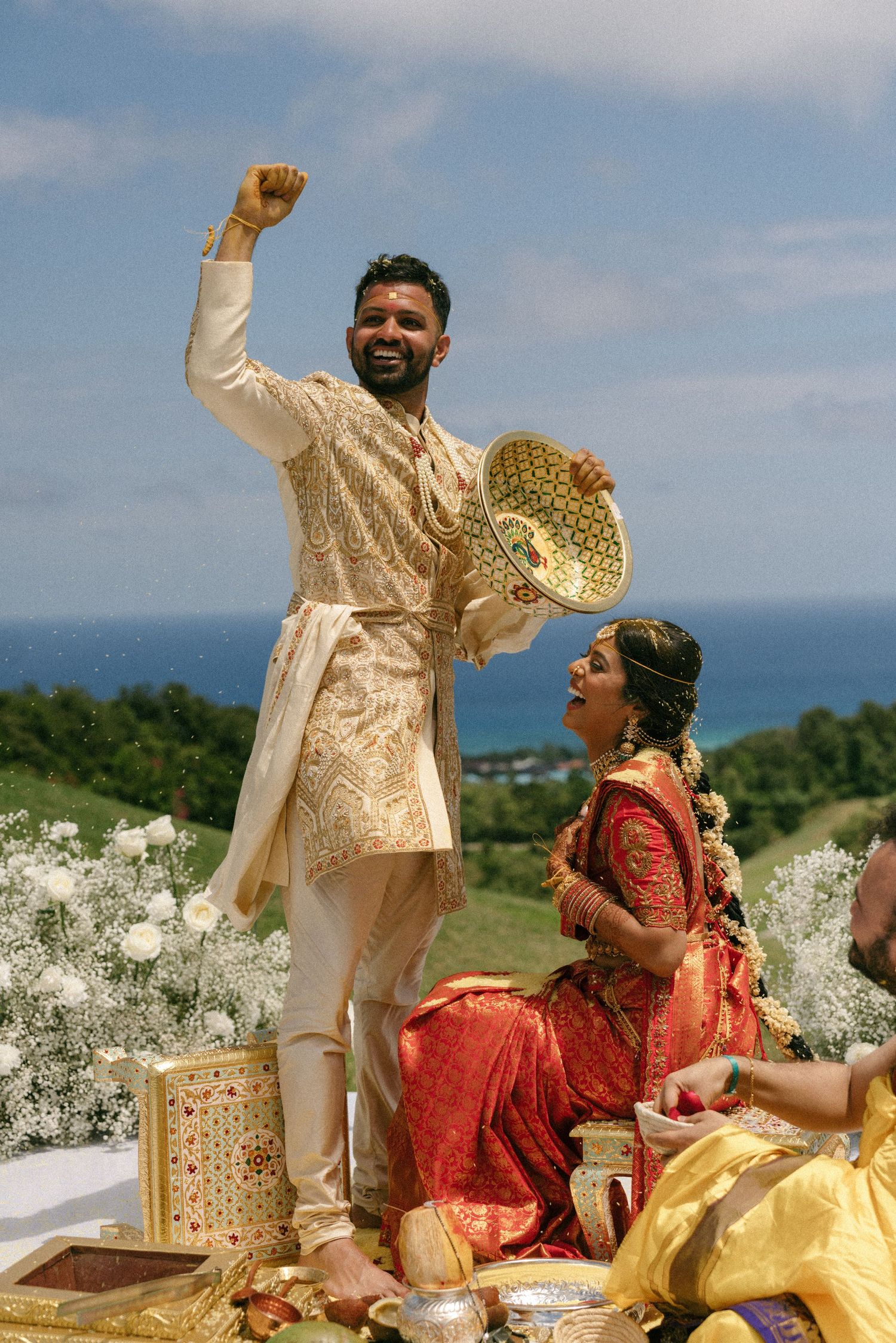 Joyful Indian groom in cream sherwani raises fist during outdoor wedding ceremony with bride in red saree by the ocean.