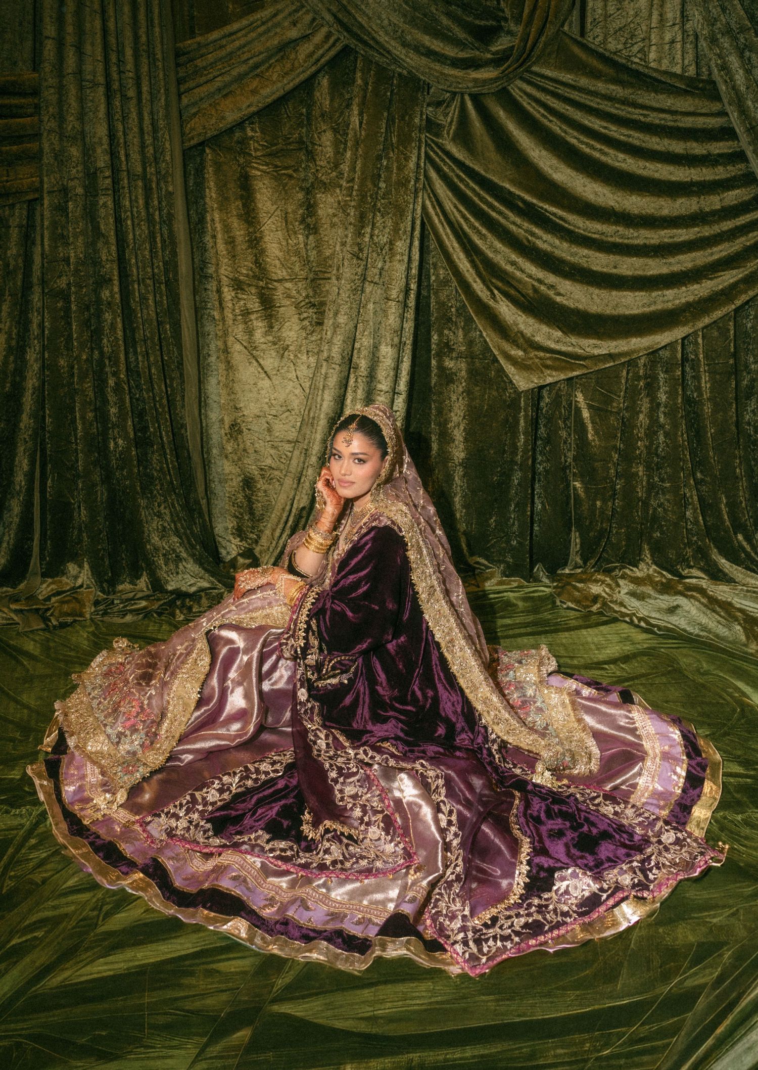 Woman in ornate purple velvet bridal lehenga with gold embroidery sits gracefully against green draped backdrop.