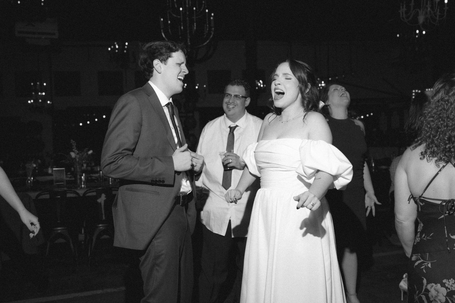 Black and white photo of a bride in white dress laughing joyfully with groom in suit at wedding reception dance floor.